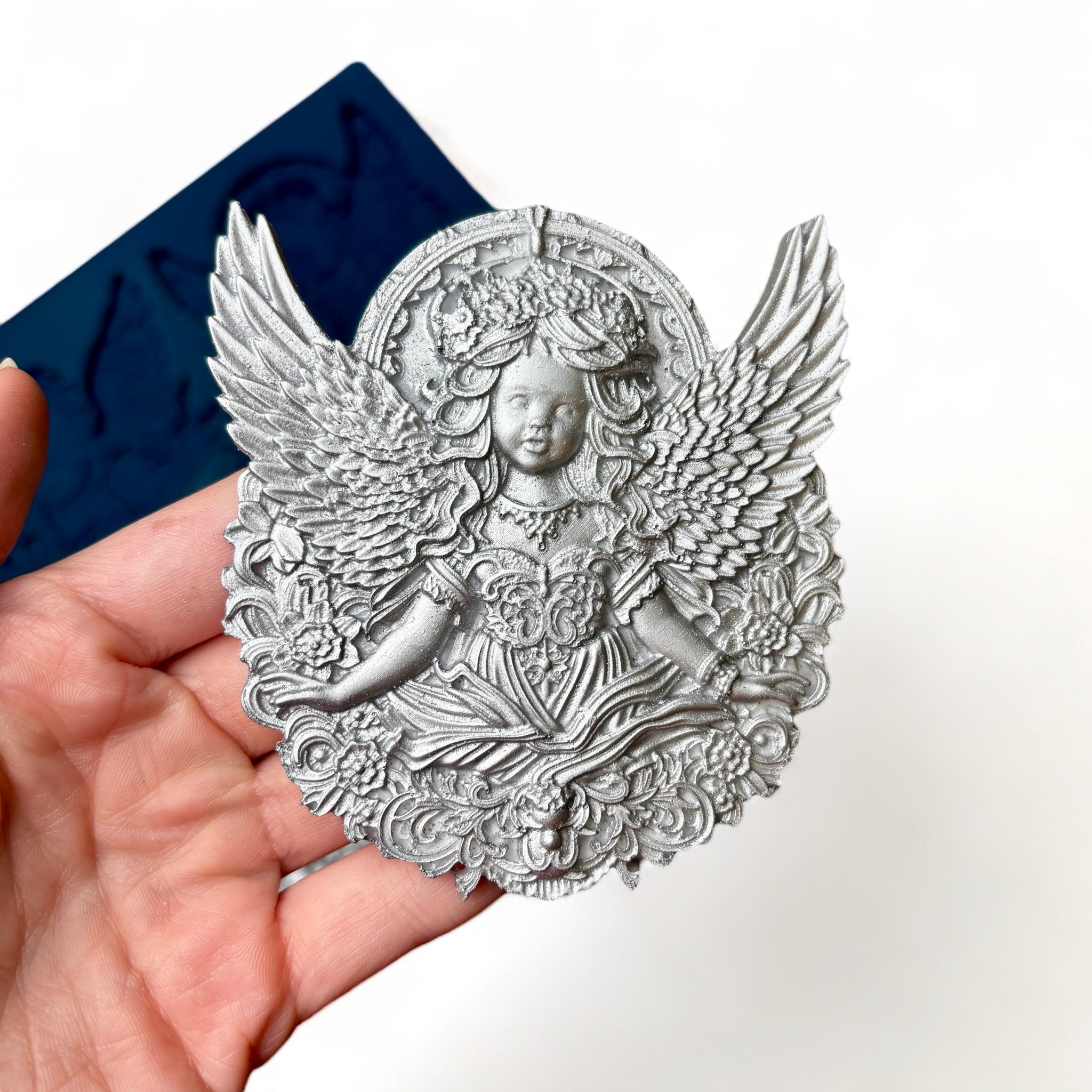 A blue silicone mold featuring 2 ornately designed cherub angels is against a white background. A hand is shown holding one of the cherub castings.