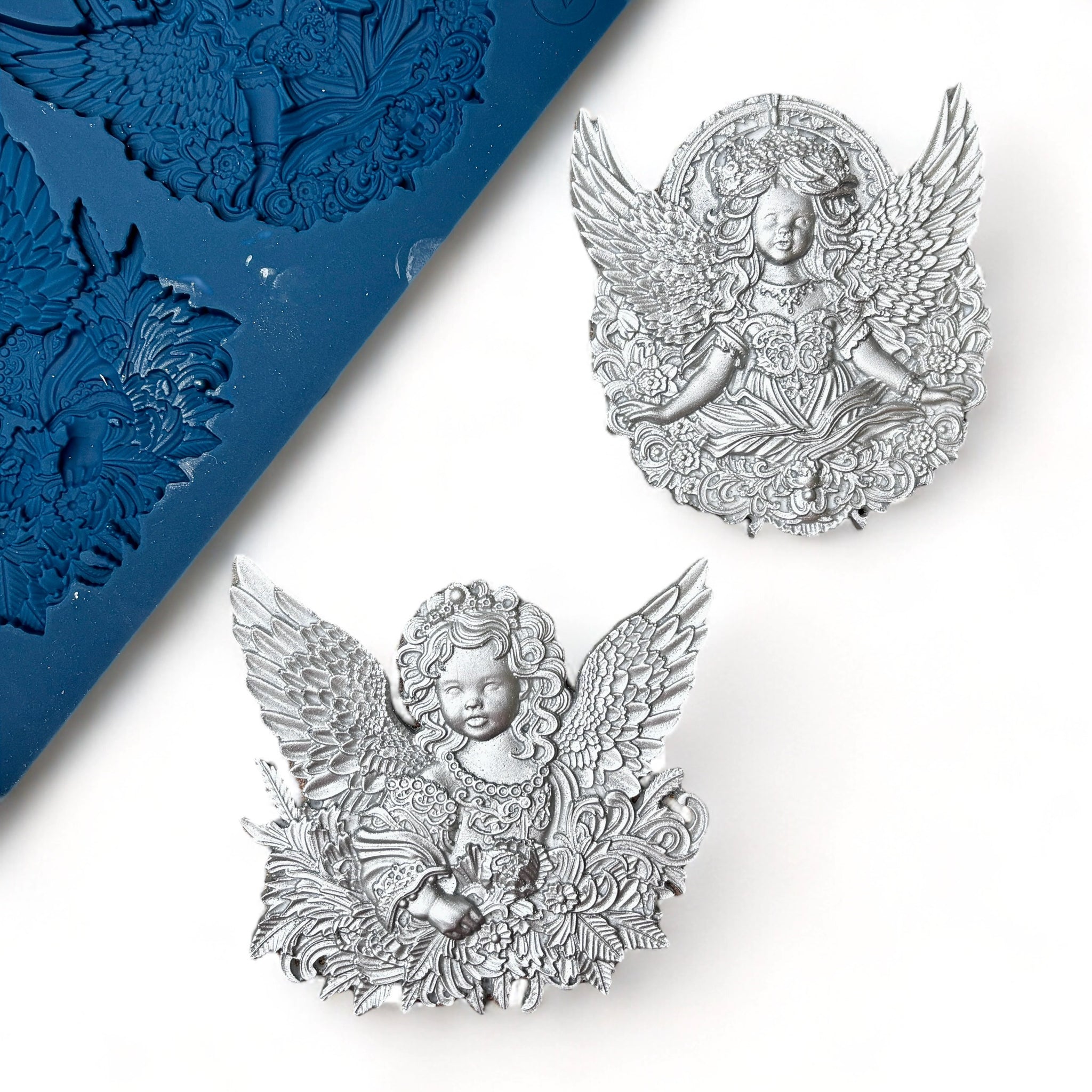 A blue silicone mold and silver colored castings of 2 ornately designed cherub angels are against a white background.