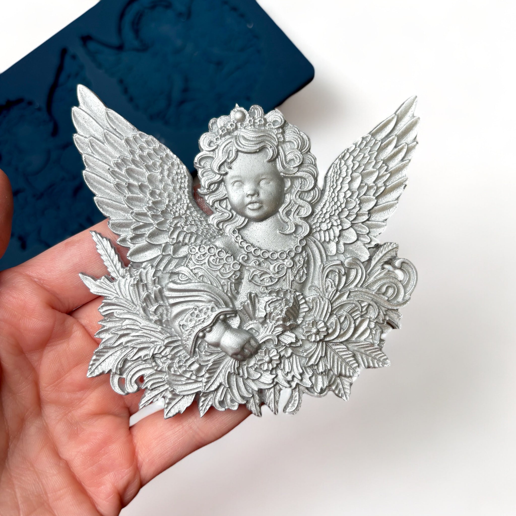 A blue silicone mold featuring 2 ornately designed cherub angels is against a white background. A hand is shown holding one of the cherub castings.