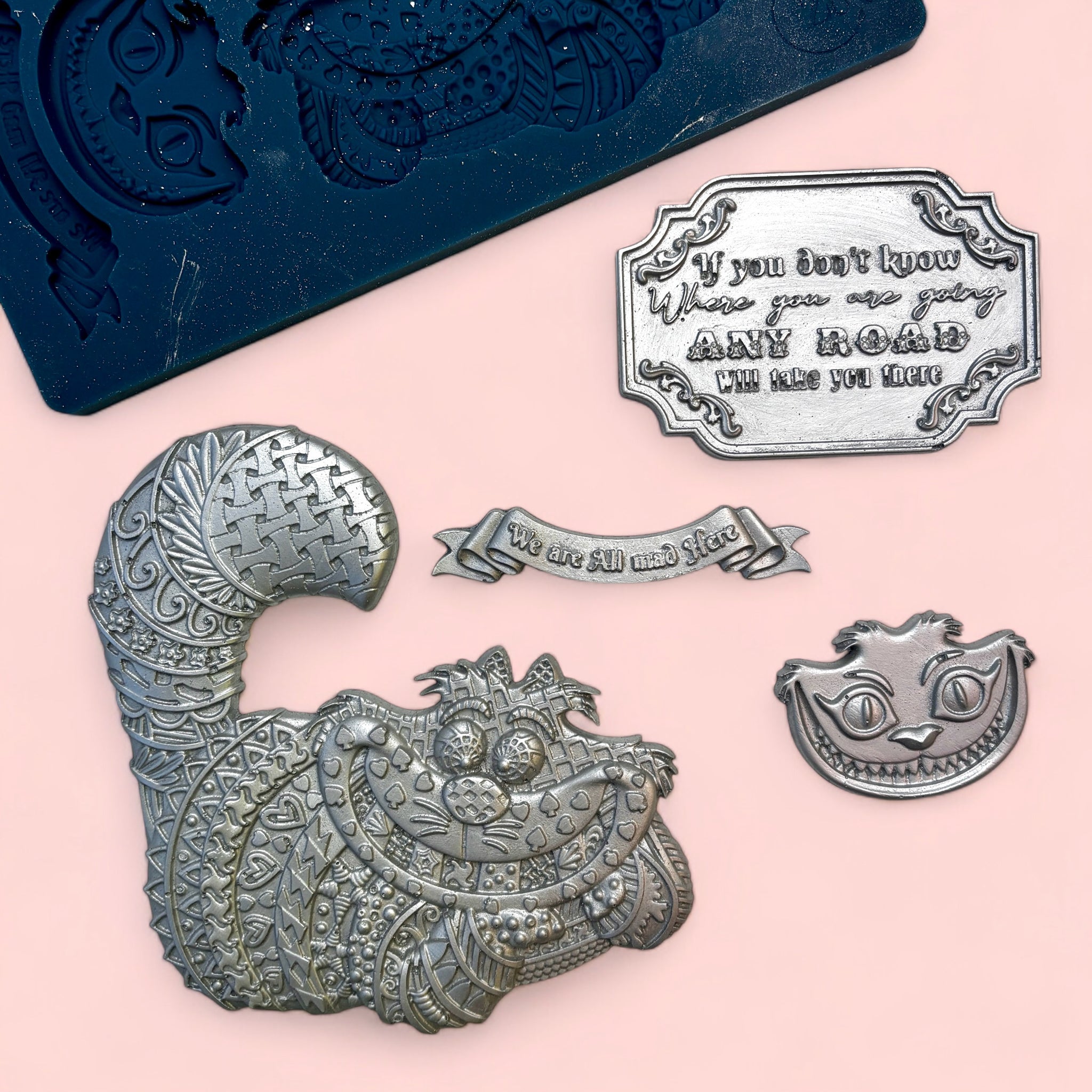 A blue silicone mold and silver colored castings of Alice in Wonderland's Cheshire cat, his face, a banner, and a sign are against a light pink background.