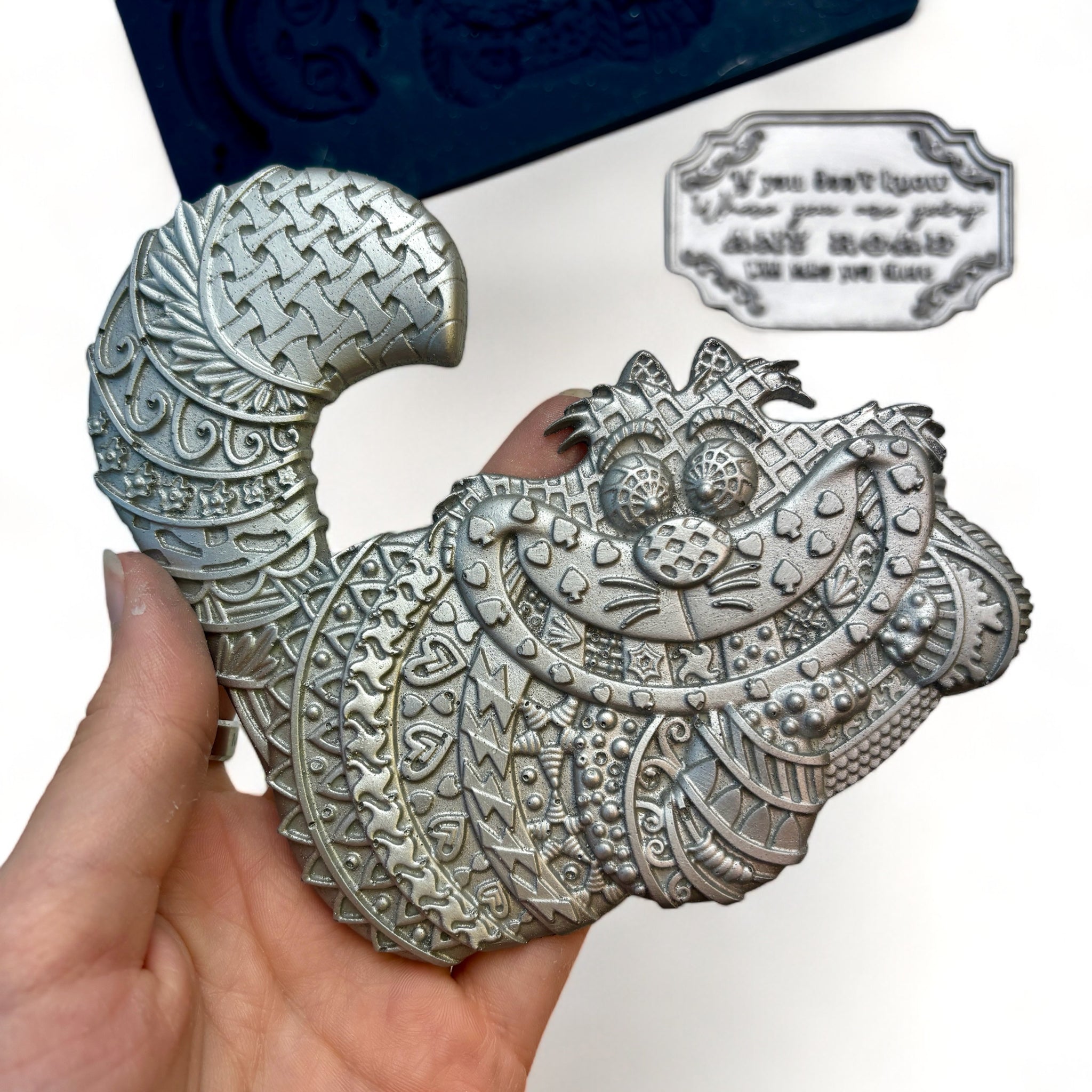 A blue silicone mold and silver colored castings of Zuri Design's Cheshire Cat are against a white background. A hand is shown holding the cat mold.