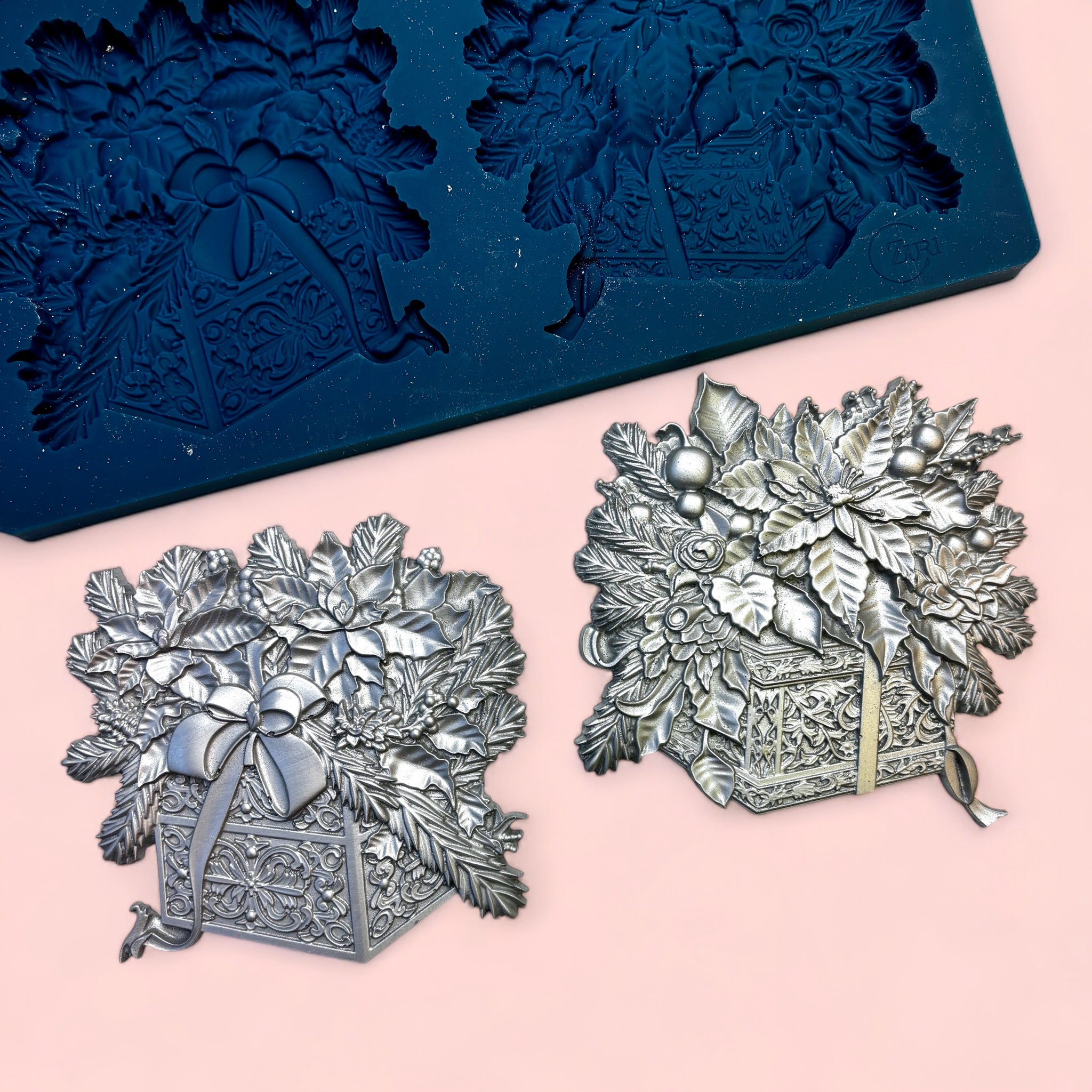 A blue silicone mold and silver colored castings of 2 holiday gifts topped with poinsettia bouquets are against a light pink background.