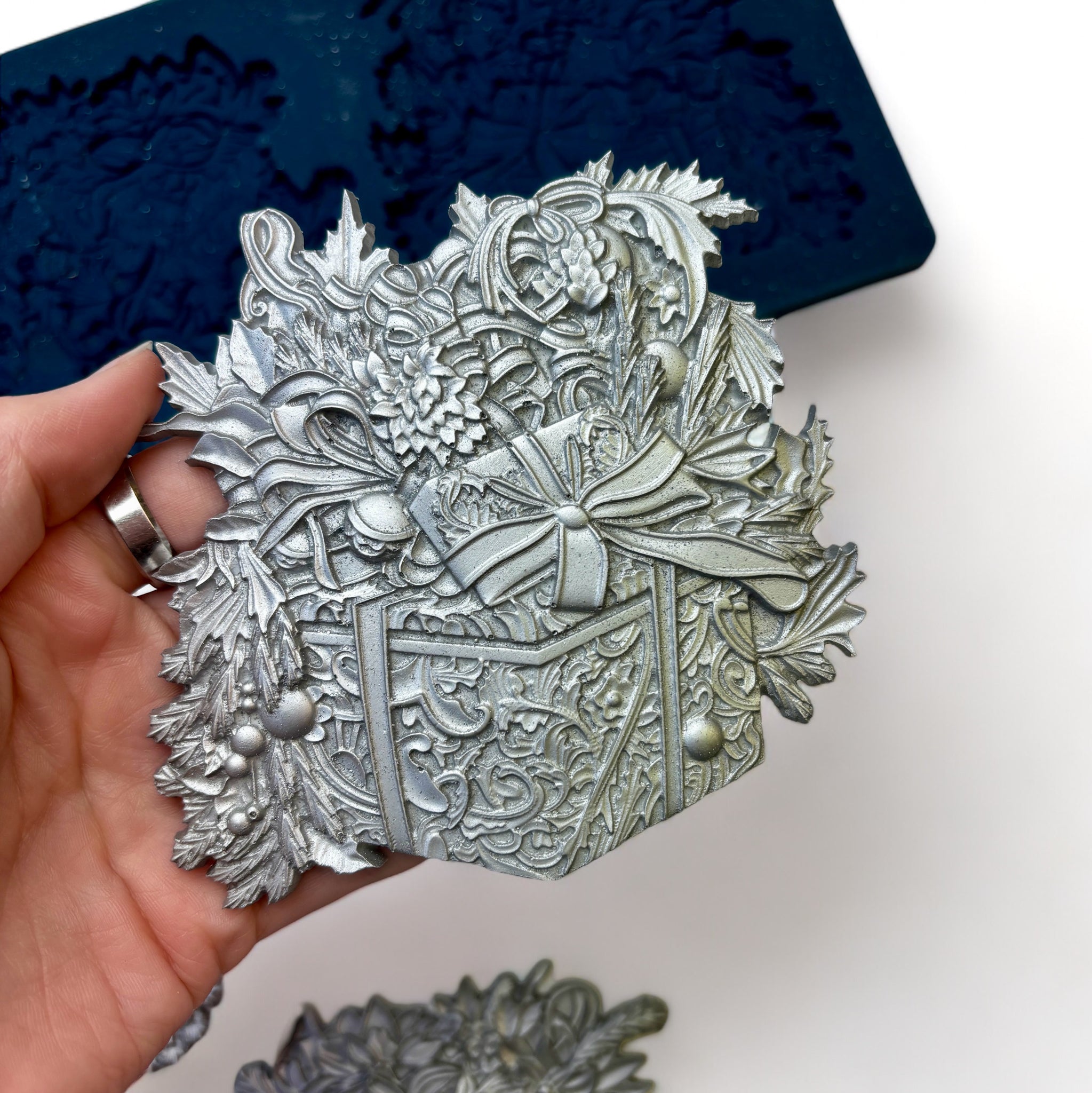 A blue silicone mold and silver colored casting of Zuri Design's Christmas Gifts Set 2 are against a white background. A hand is shown holding one of the castings.