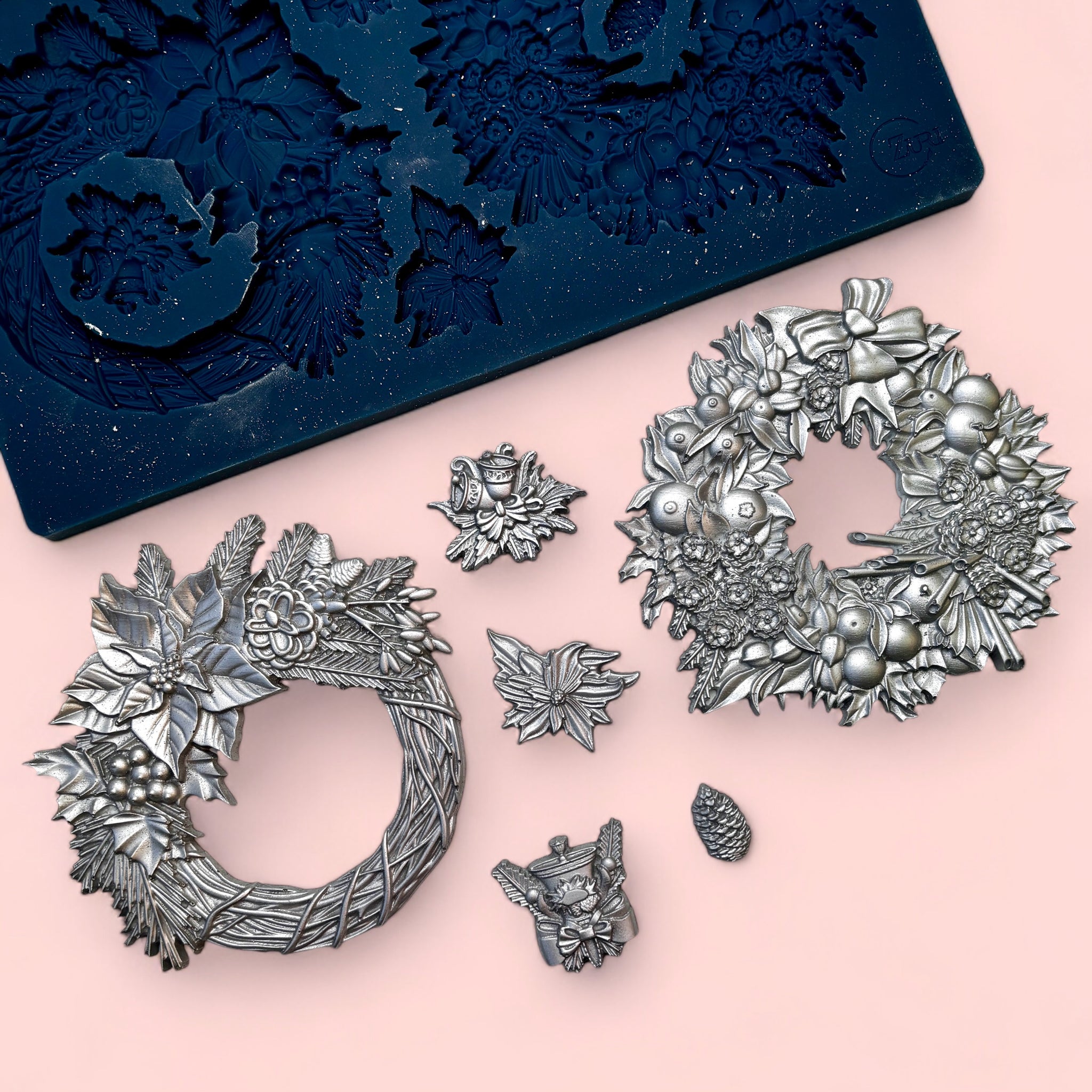 A blue silicone mold and silver colored castings of 2 holiday wreaths and 4 small accent pieces are against a light pink background.