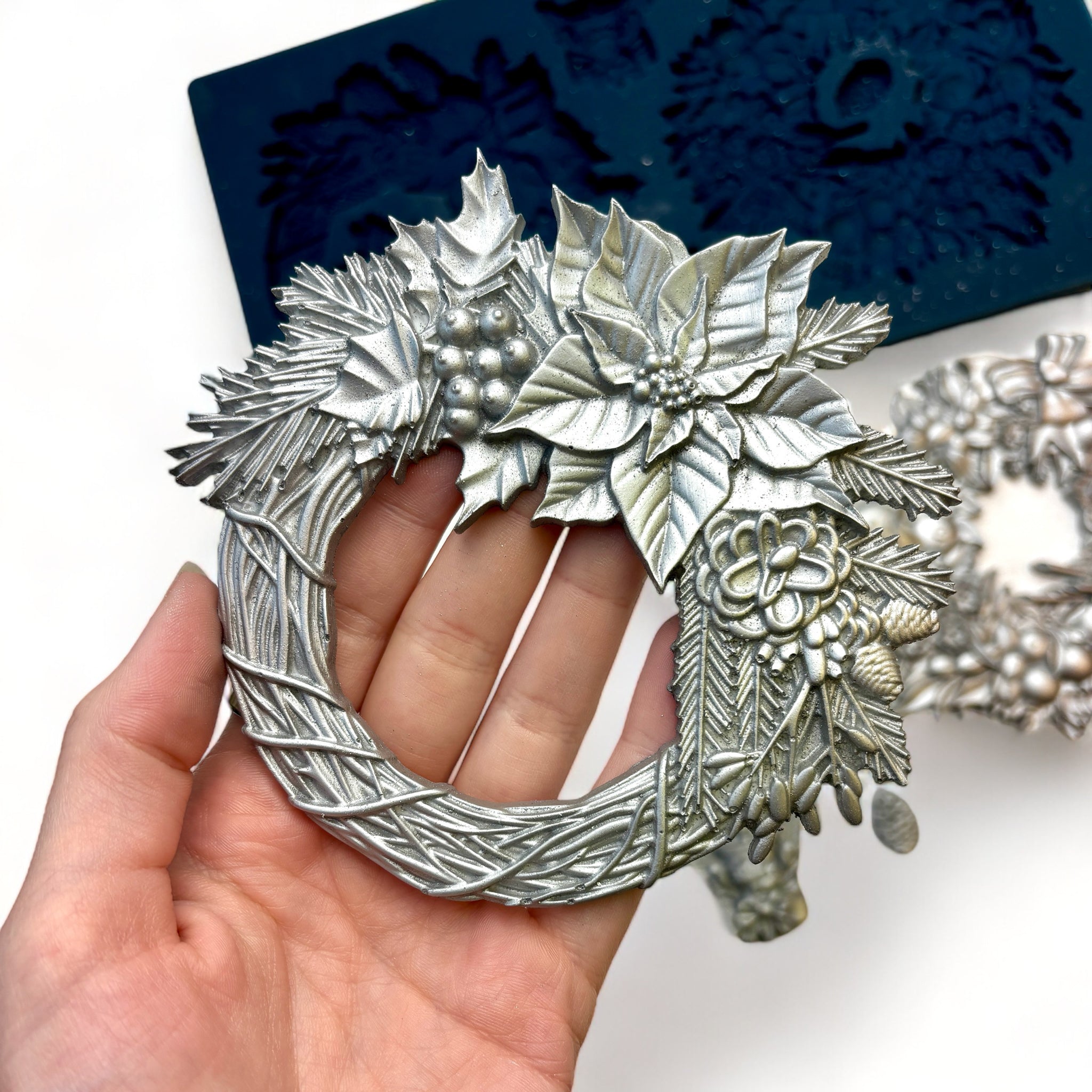 A blue silicone mold and silver colored castings of Zuri Design's Christmas Wreaths are against a white background. A hand is shown holding one of the wreath castings.