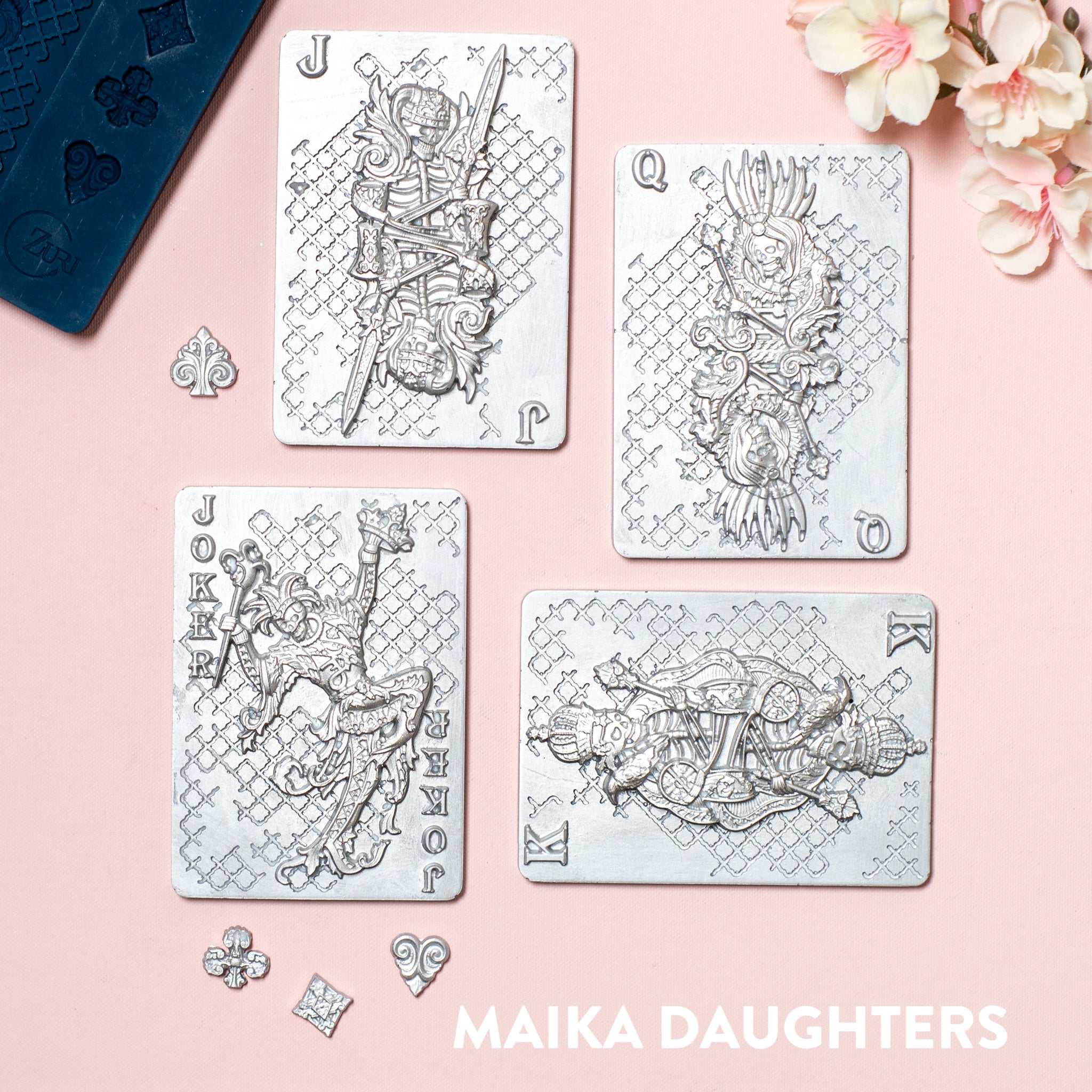 A blue silicone mold and silver colored castings of 4 playing cards that feature a king, queen, jack, and joker with skeleton face designs are against a light pink background.