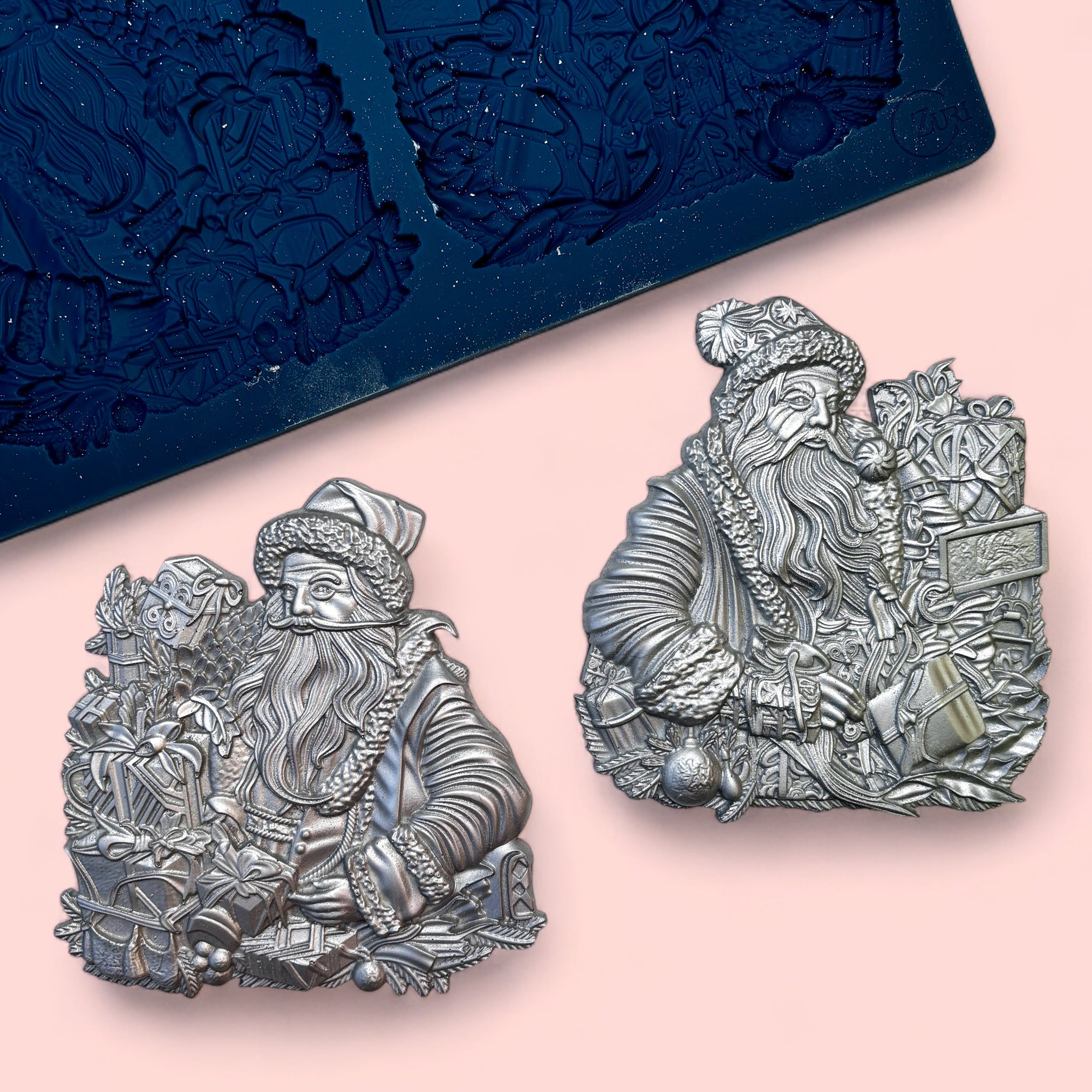 A blue silicone mold and silver colored castings of 2 Santas with stack of gifts are against a light pink background. 