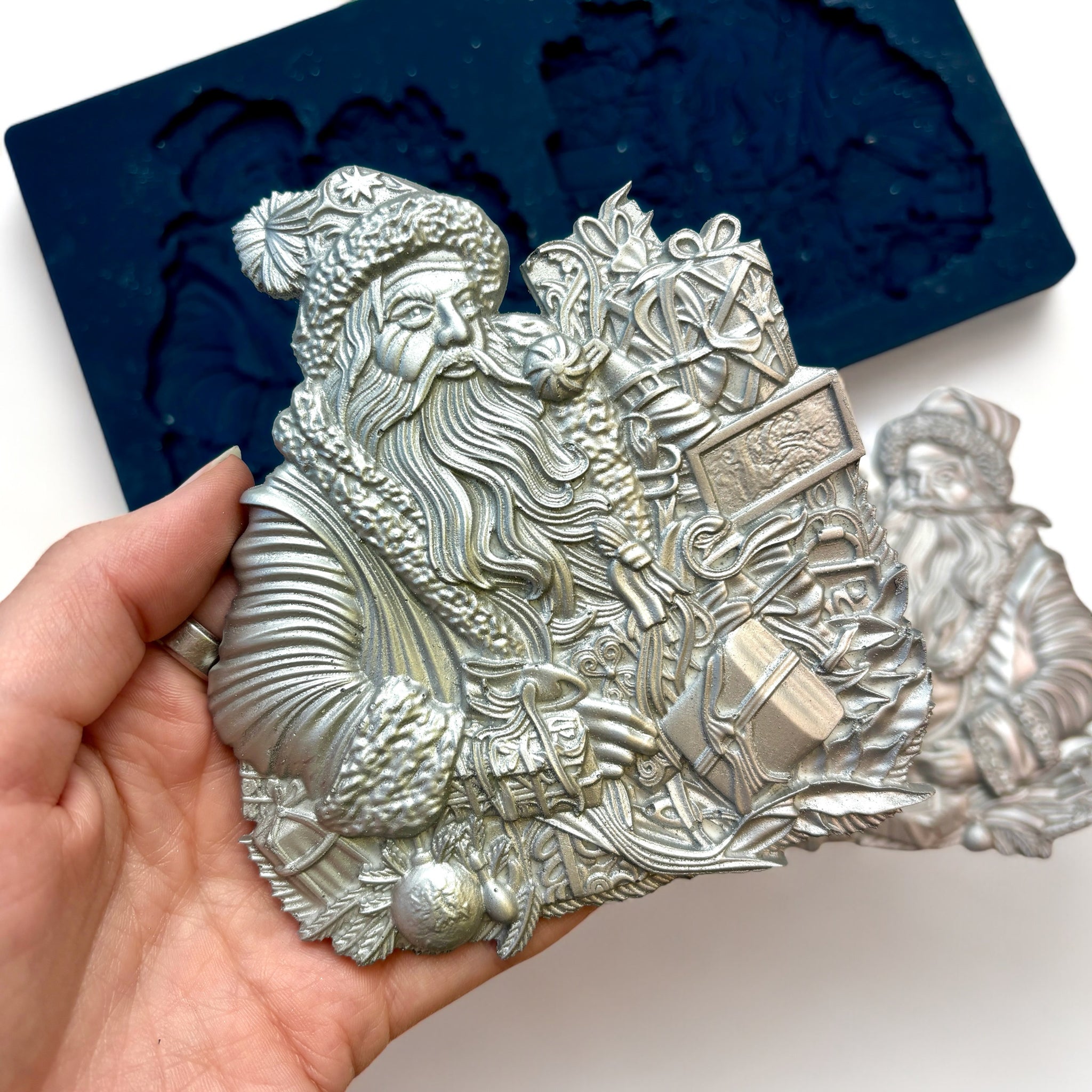 A blue silicone mold and silver colored castings of Zuri Design's Father Christmas are against a white background. A hand is shown holding one of the castings.