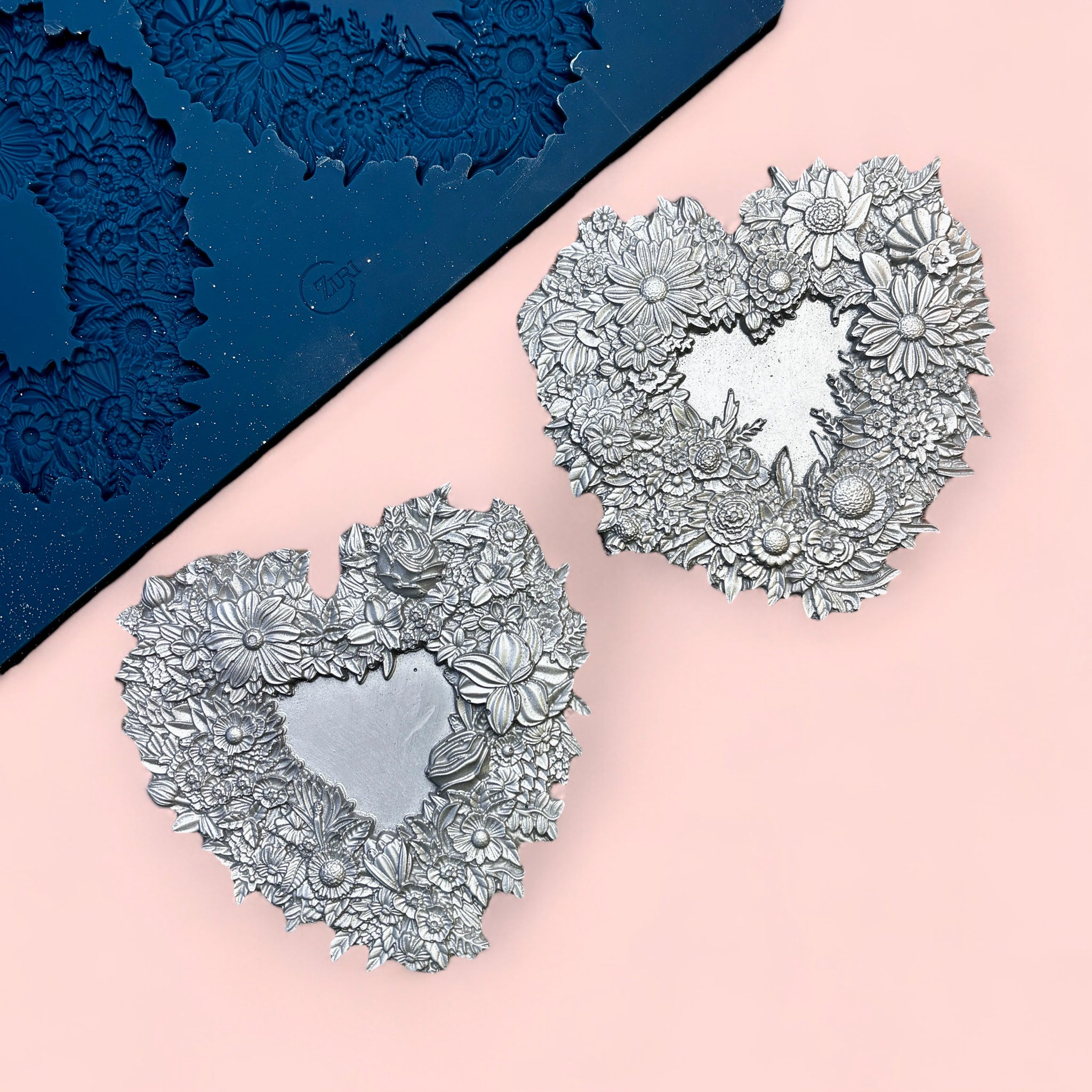 A blue silicone mold and silver colored castings of 2 floral heart frames are against a light pink background.