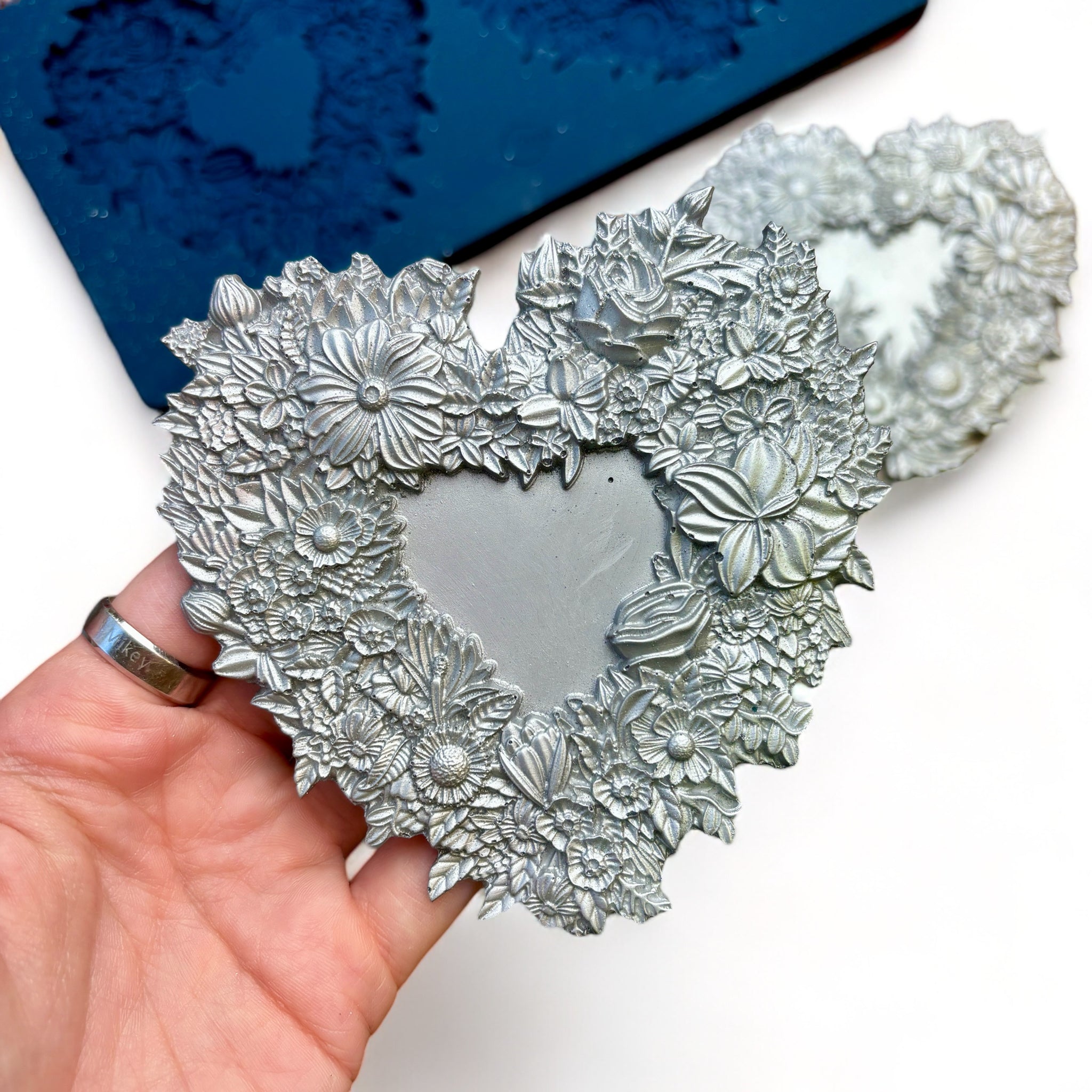 A blue silicone mold and silver colored castings of 2 floral heart frames are against a white background. A hand is shown holding one of the castings.