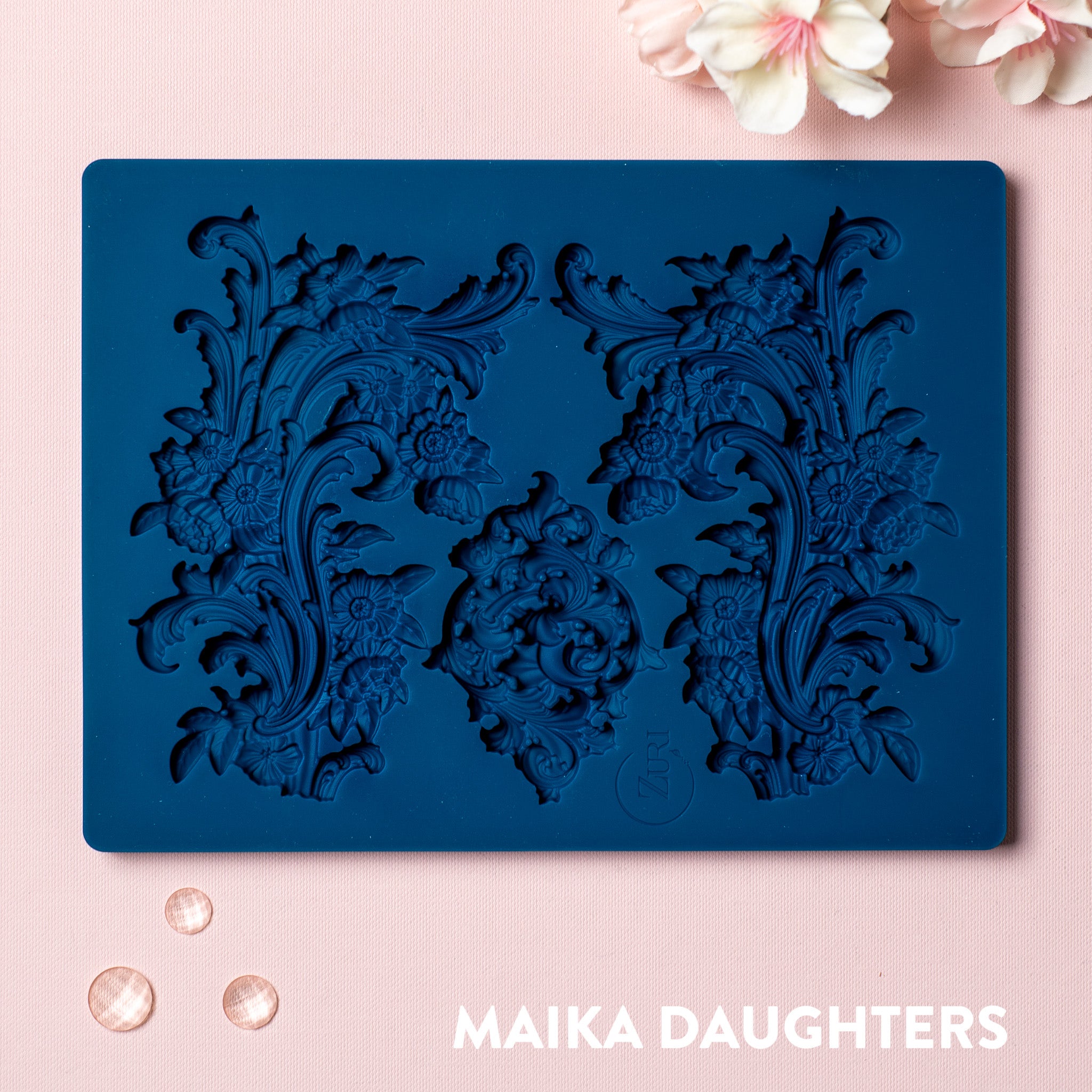 A blue silicone mold featuring 3 ornate floral scroll flourish accent pieces is against a light pink background.