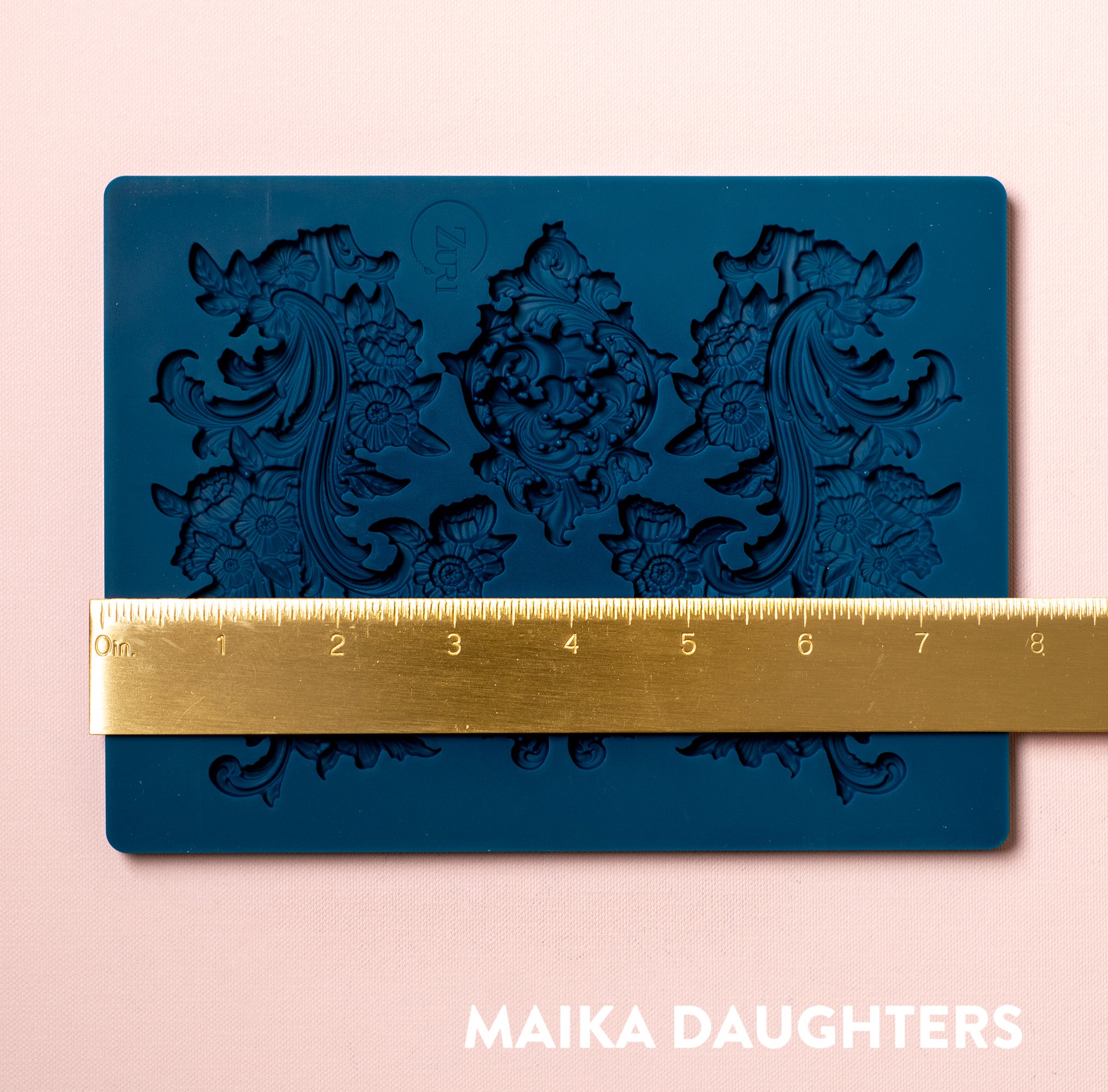 A blue silicone mold featuring 3 ornate floral scroll flourish accent pieces is against a light pink background. A gold ruler reading 7.8" width sits on the mold.