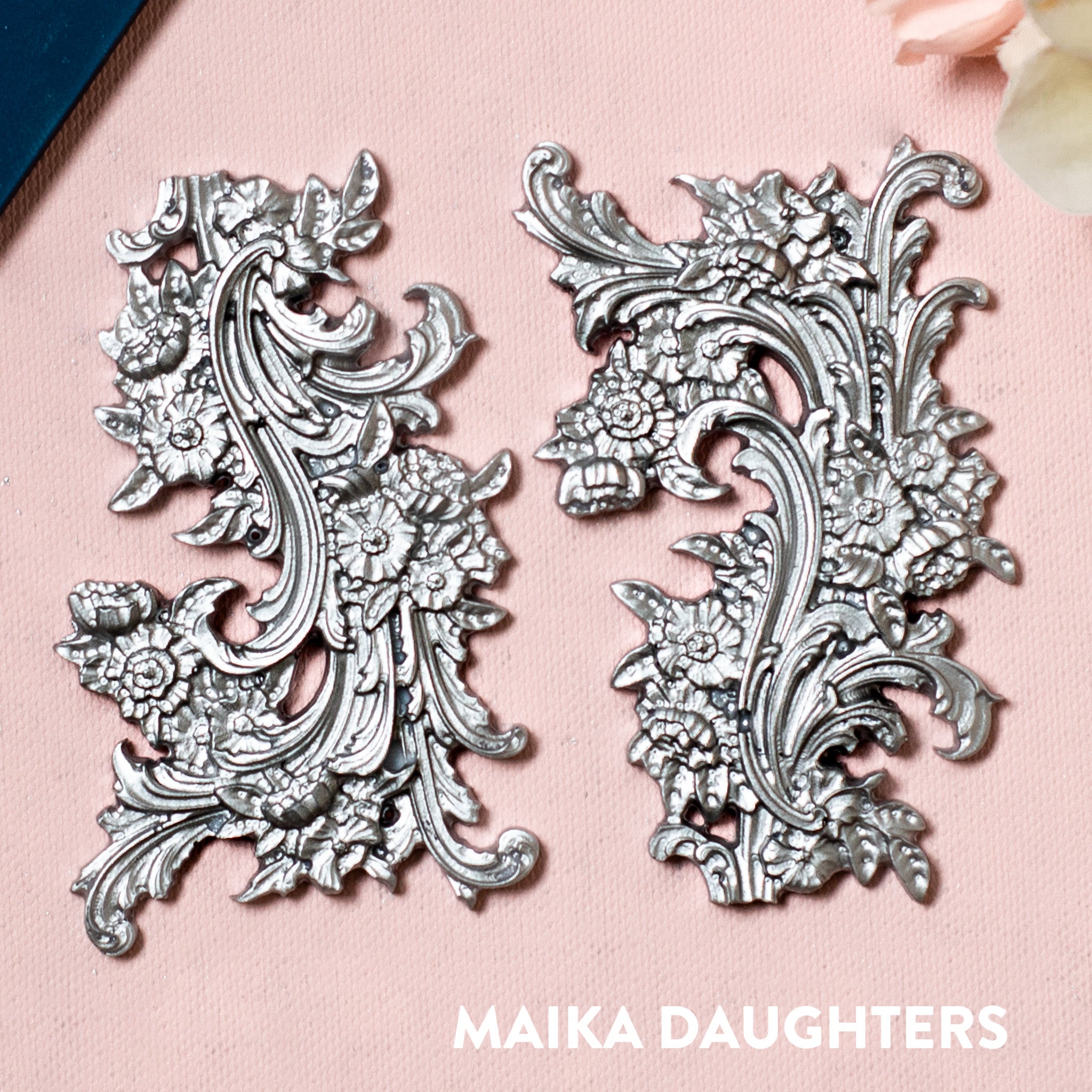 Silver colored silicone mold castings featuring 2 small ornate floral flourish accent pieces are against a pink background. 