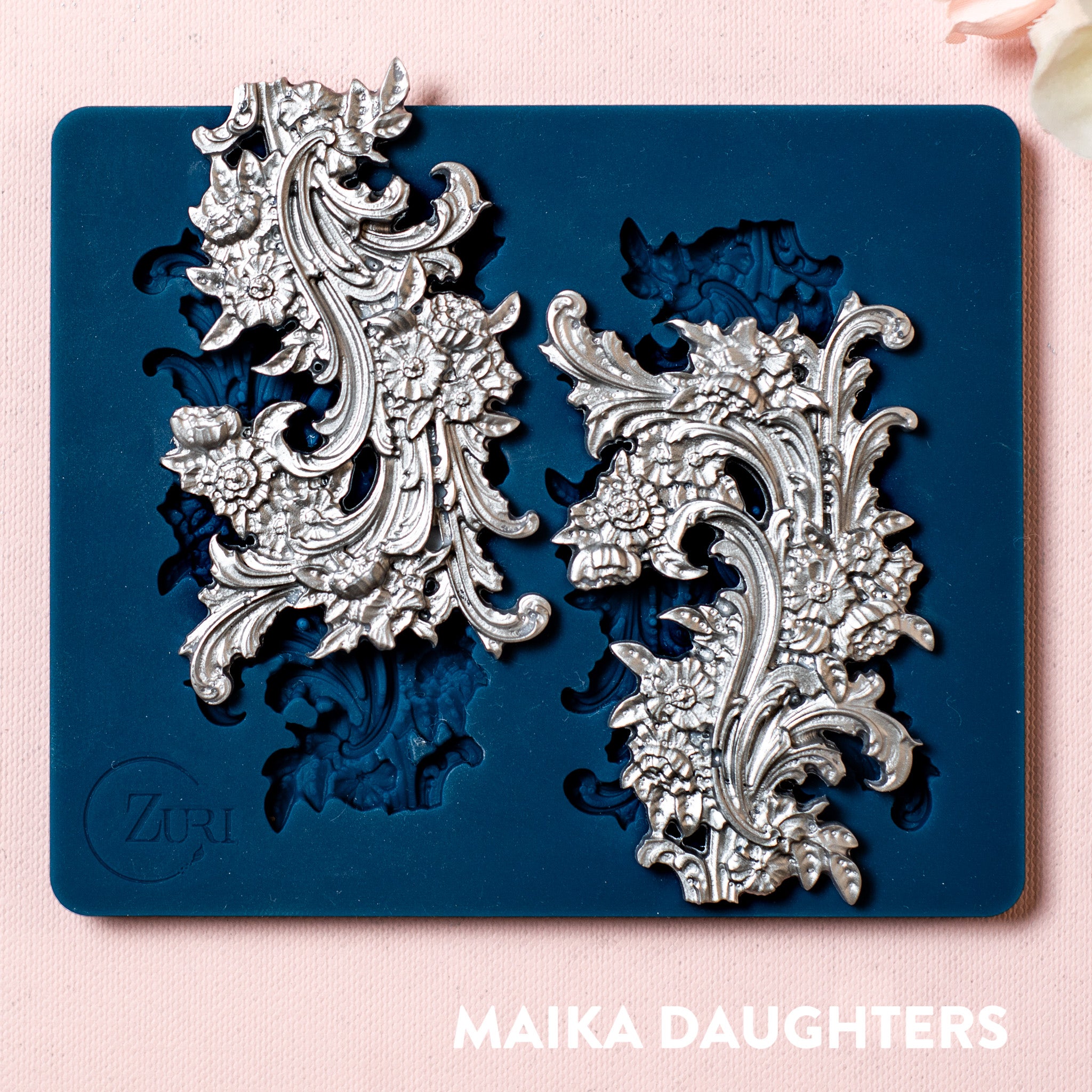 A blue silicone mold and silver colored castings of Zuri Design's Flourishes Set 1 small are against a light pink background.