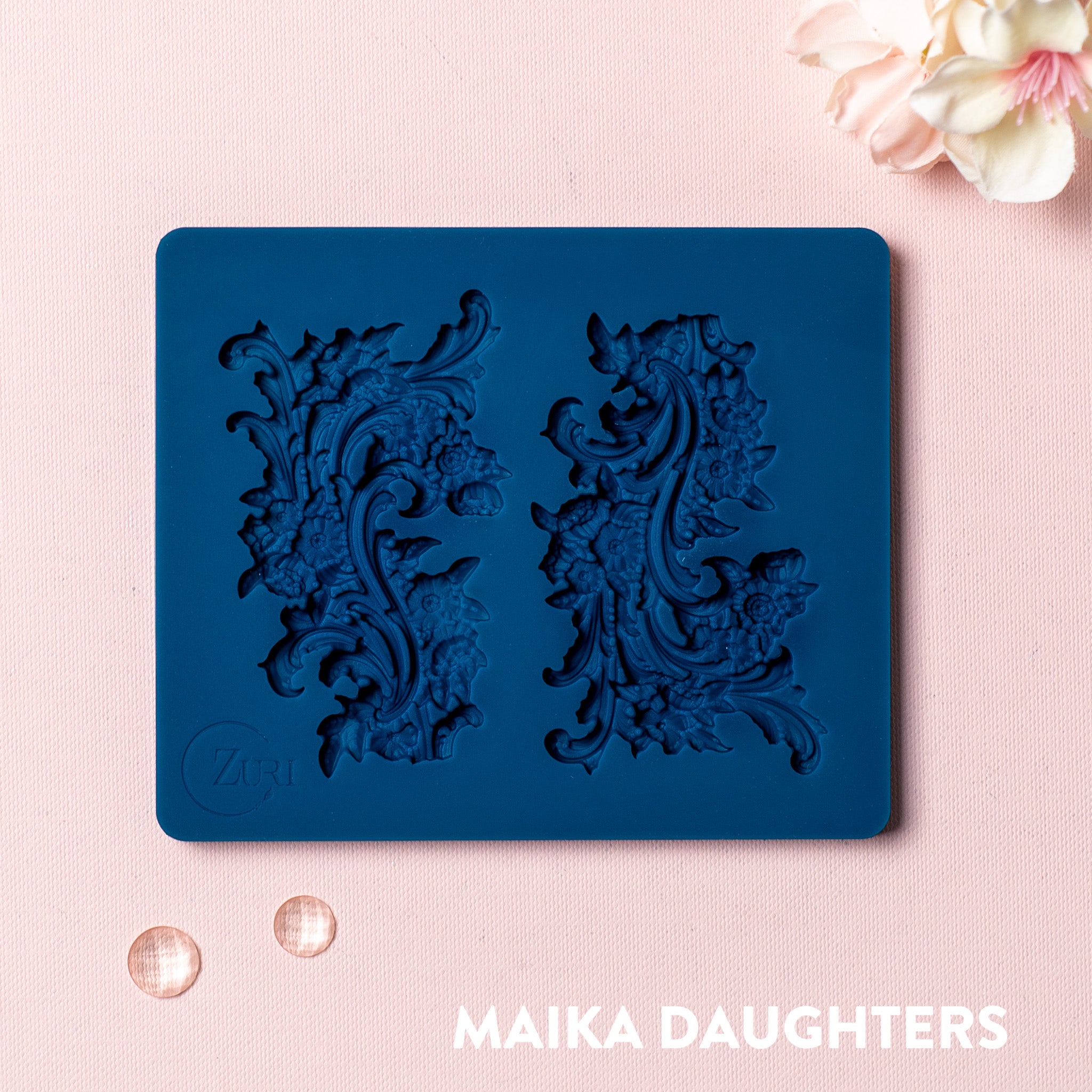 A blue silicone mold featuring 2 small ornate floral flourish accent pieces is against a light pink background.