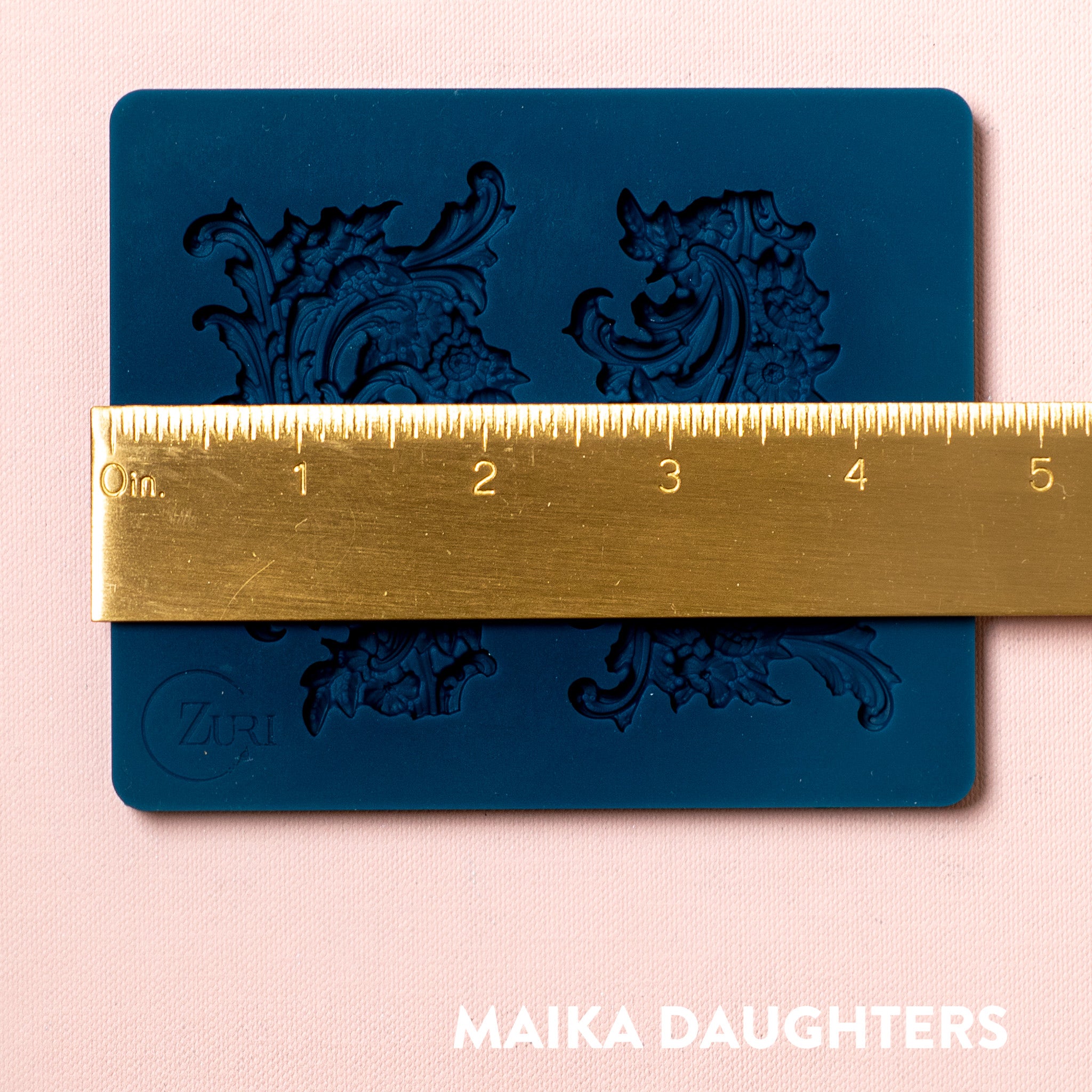 A blue silicone mold featuring 2 small ornate floral flourish accent pieces is against a light pink background. A gold ruler reading 4.7" width sits on the mold.