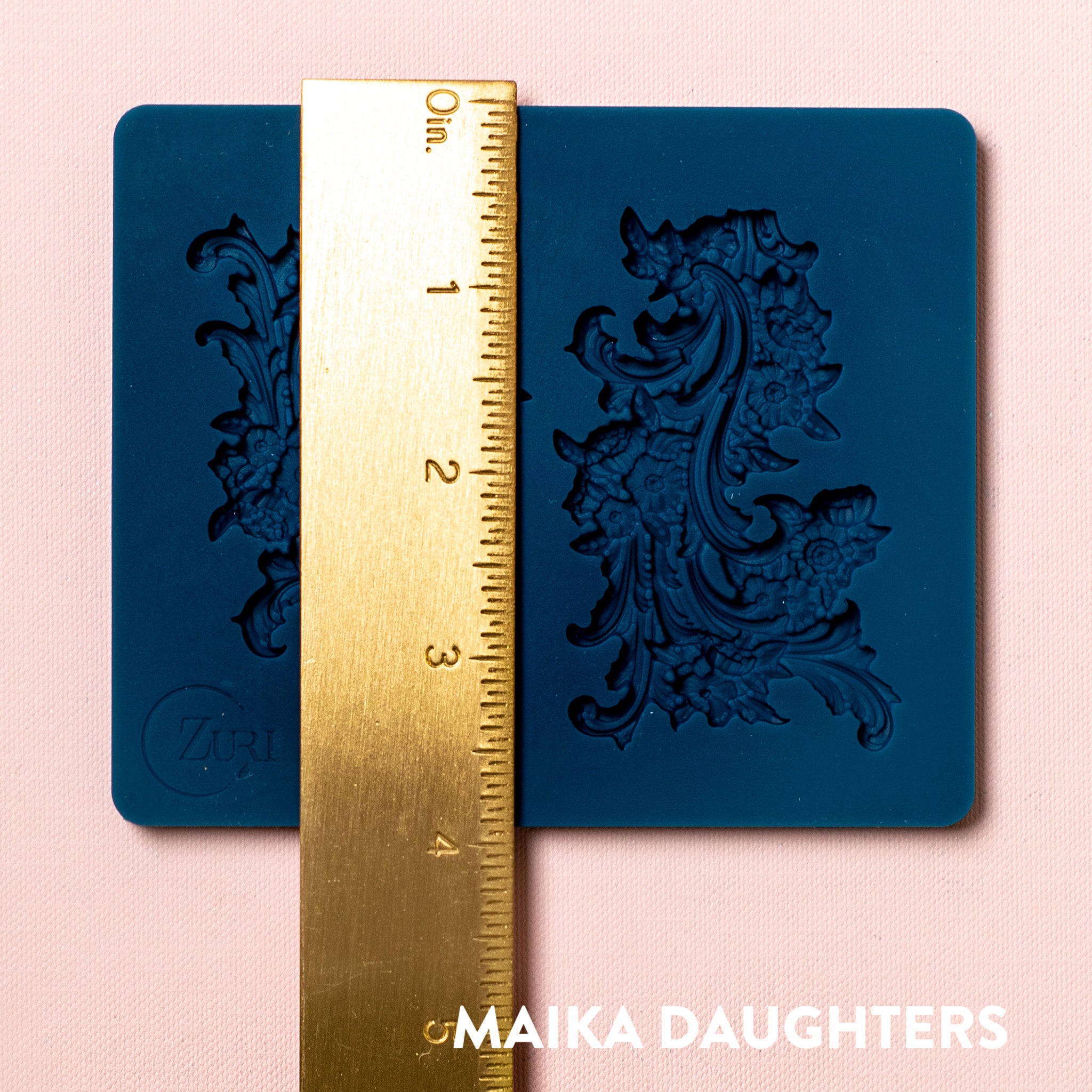 A blue silicone mold featuring 2 small ornate floral flourish accent pieces is against a light pink background. A gold ruler reading 3.9" height sits on the mold.