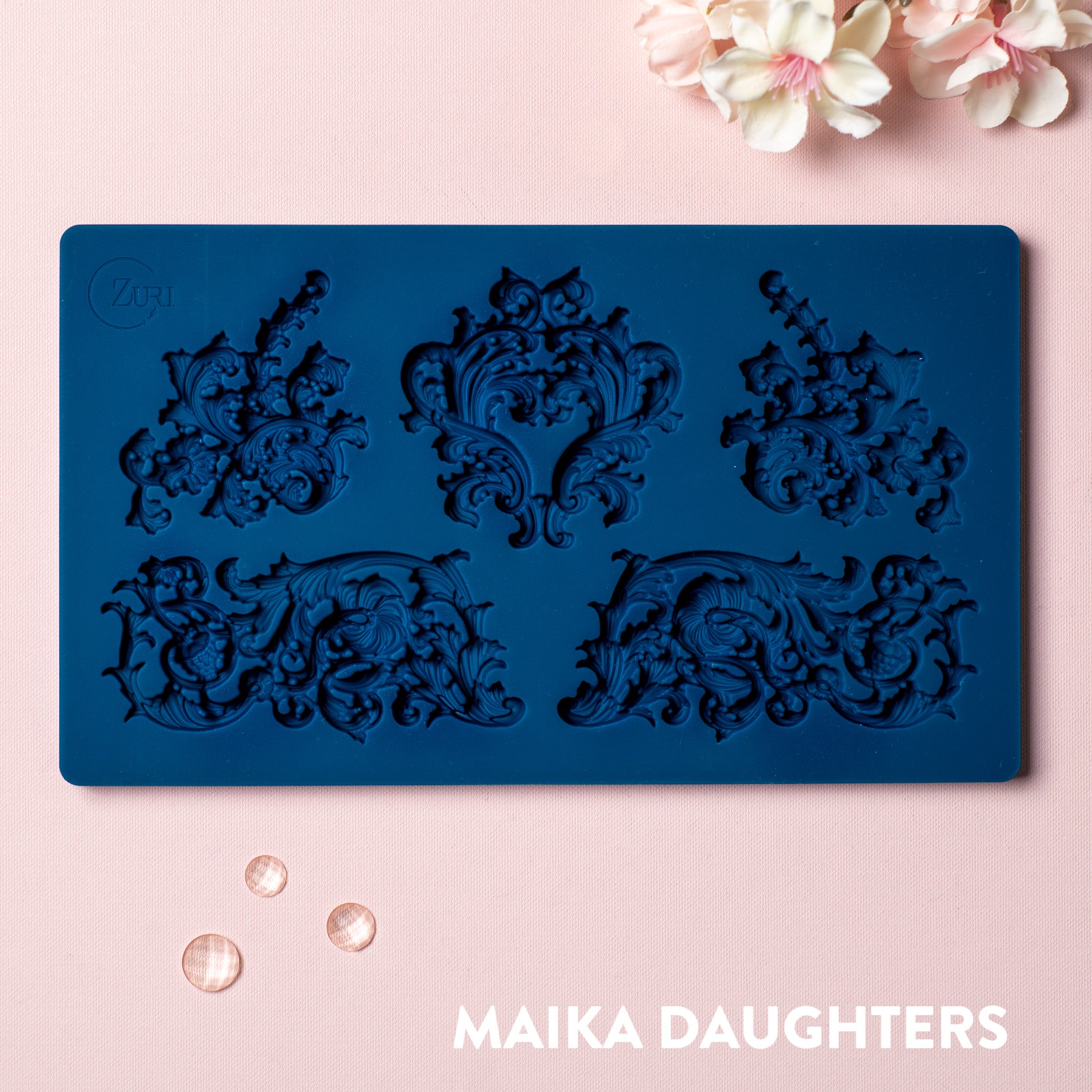 A blue silicone mold featuring 5 ornate flourish accent pieces is against a light pink background.