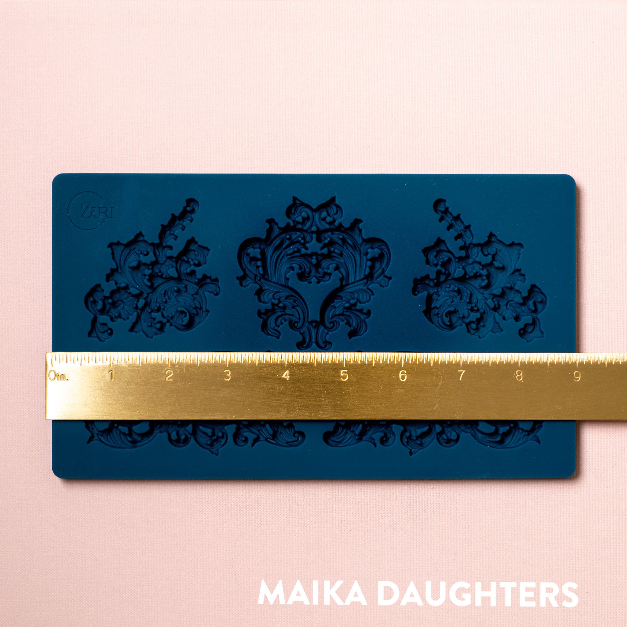 A blue silicone mold featuring 5 ornate flourish accent pieces is against a light pink background. A gold ruler reading 9" width sits on the mold.