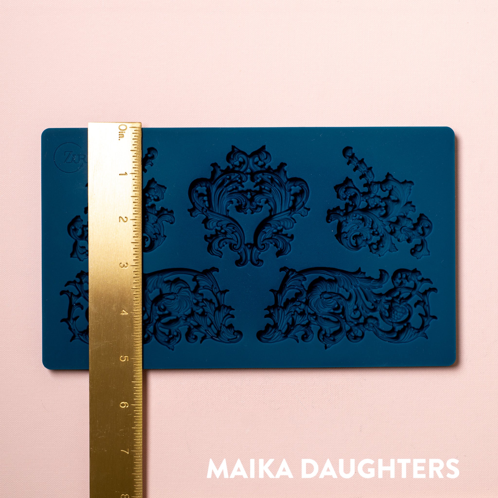 A blue silicone mold featuring 5 ornate flourish accent pieces is against a light pink background. A gold ruler reading 5.25" height sits on the mold.