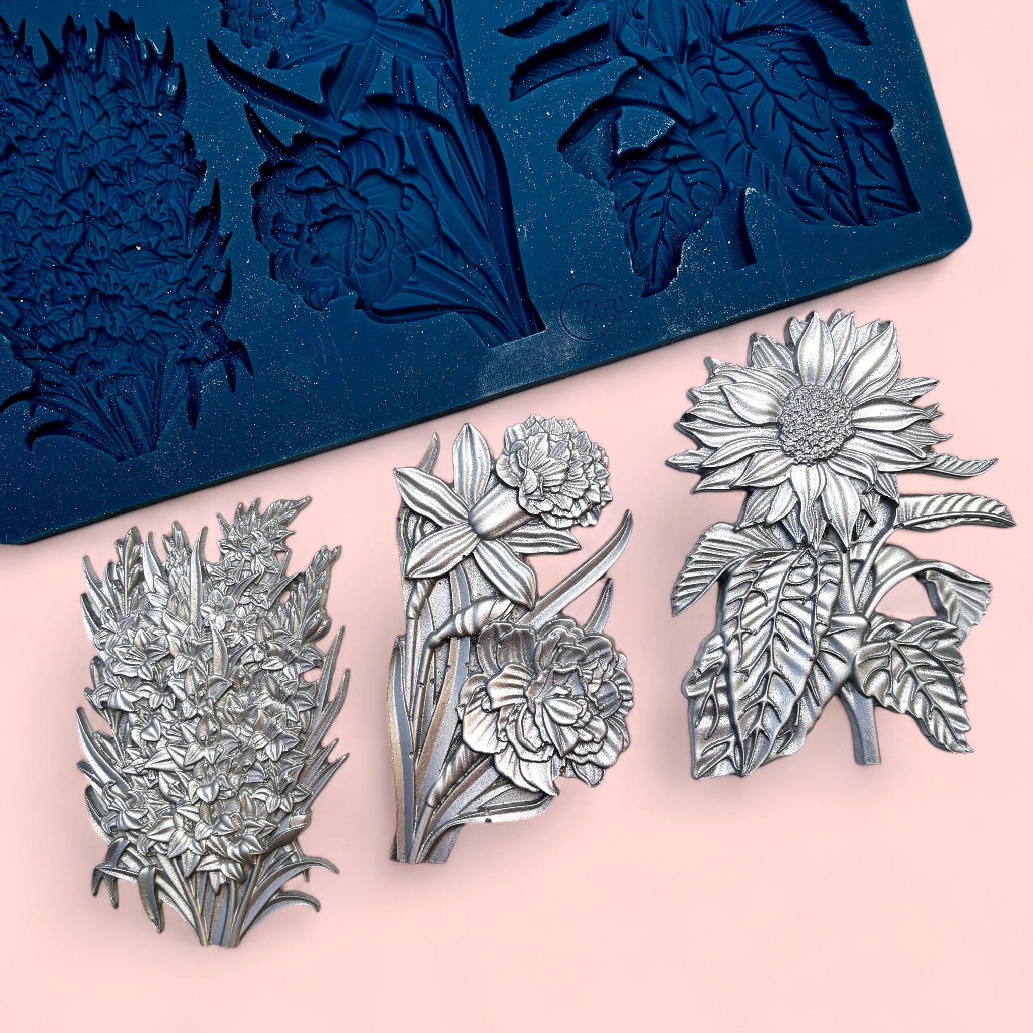 A blue silicone mold and silver colored castings of 3 clusters of flowers are against a light pink background.