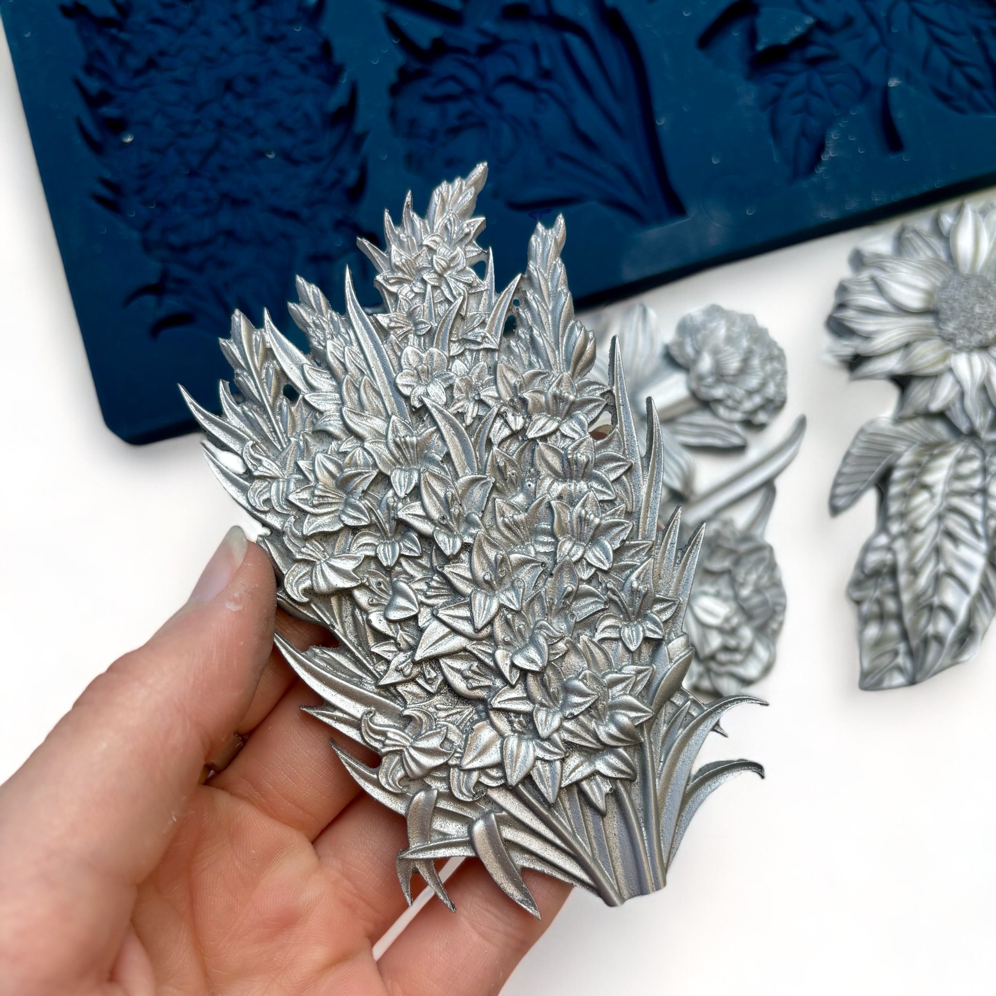 A blue silicone mold and silver colored castings of Zuri Design's Flower Blooms Set 1 are against a white background. A hand is shown holding one of the castings.