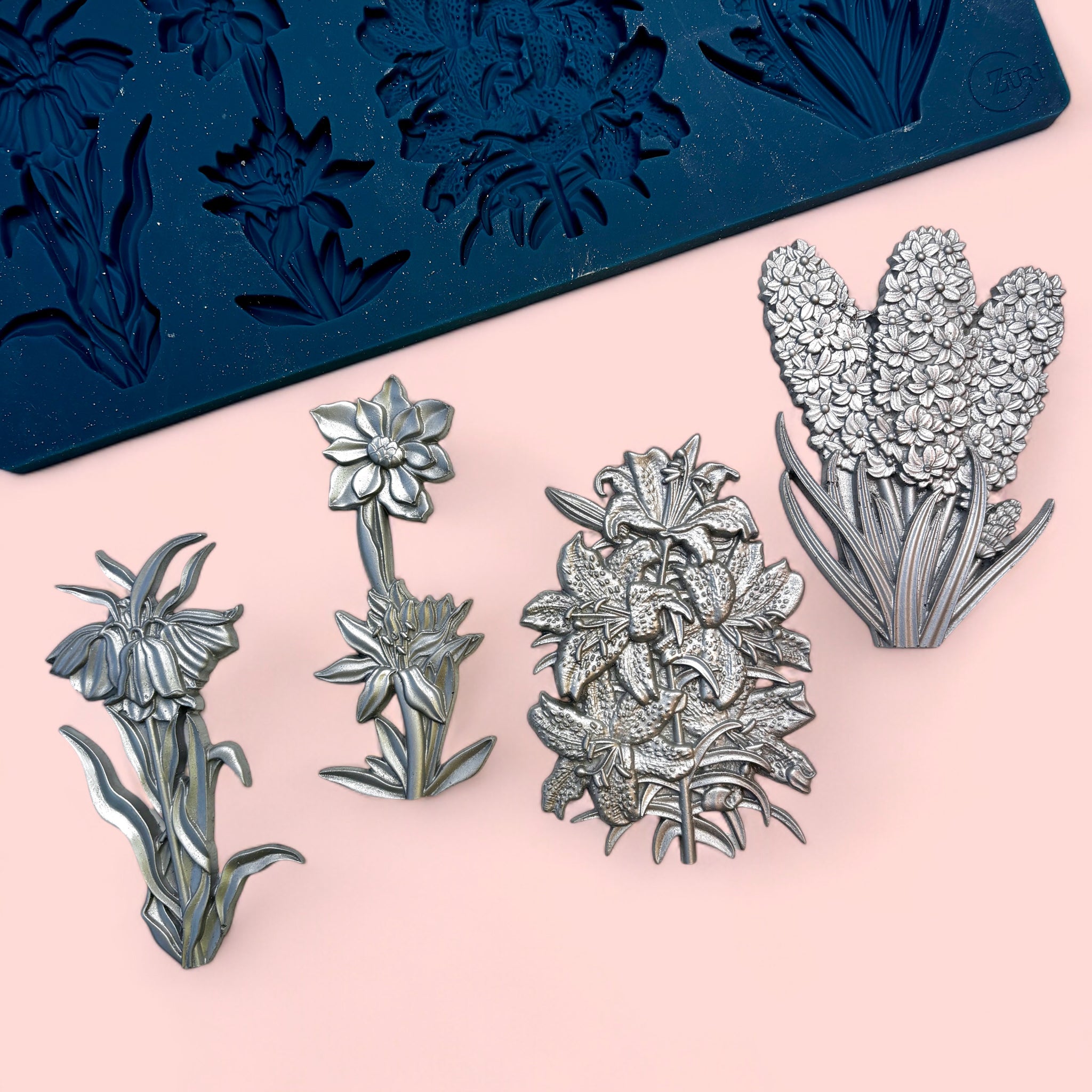 A blue silicone mold and silver colored castings of 4 clusters of flowers are against a light pink background.