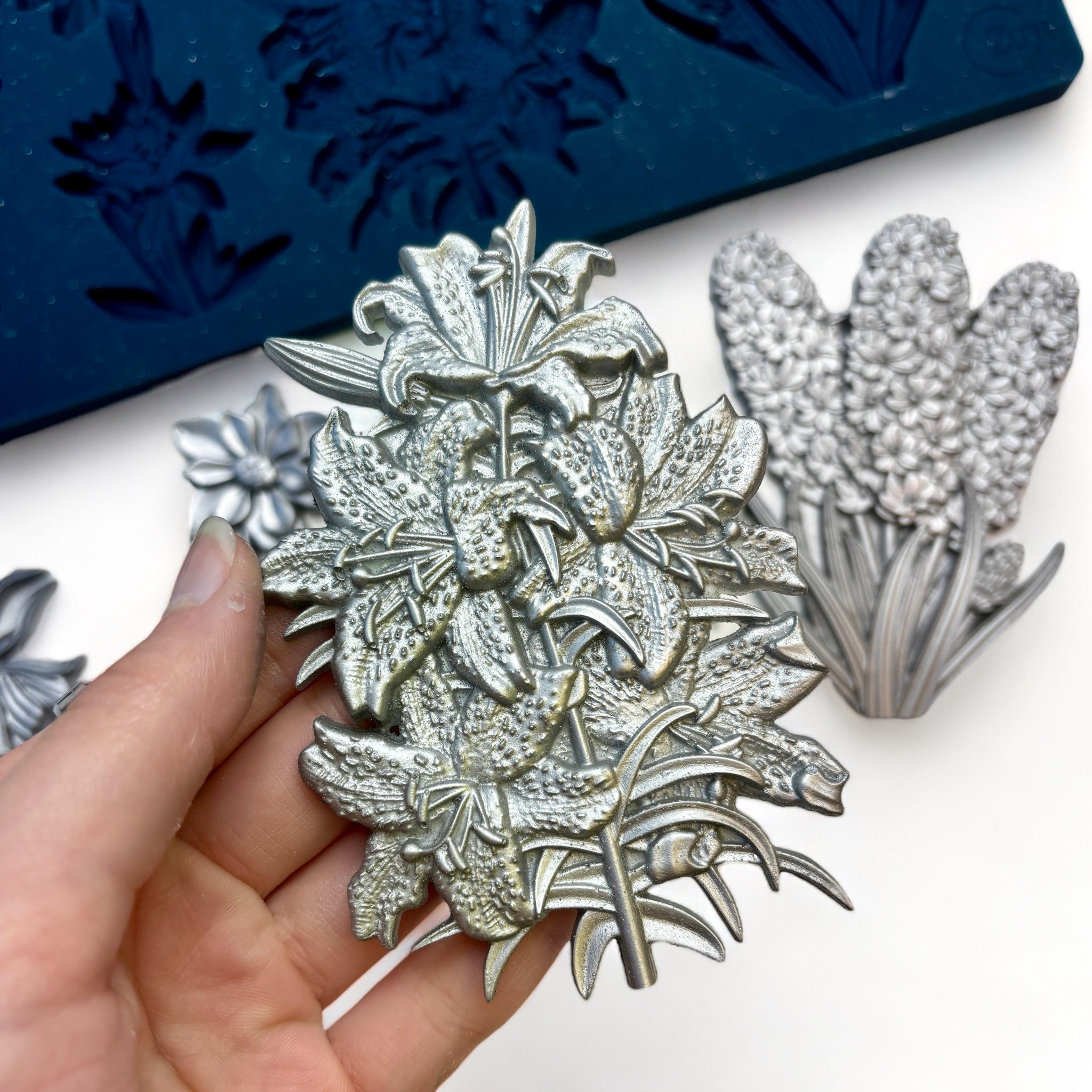 A blue silicone mold and silver colored castings of Zuri Design's Flower Blooms Set 2 are against a white background. A hand is shown holding one of the castings.