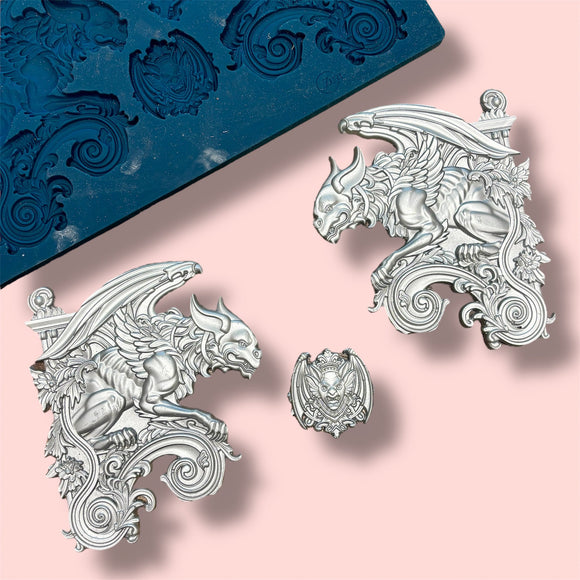 A blue silicone mold and silver colored castings featuring 2 large gargoyle statues and a smaller gargoyle medallion are against a light pink background.