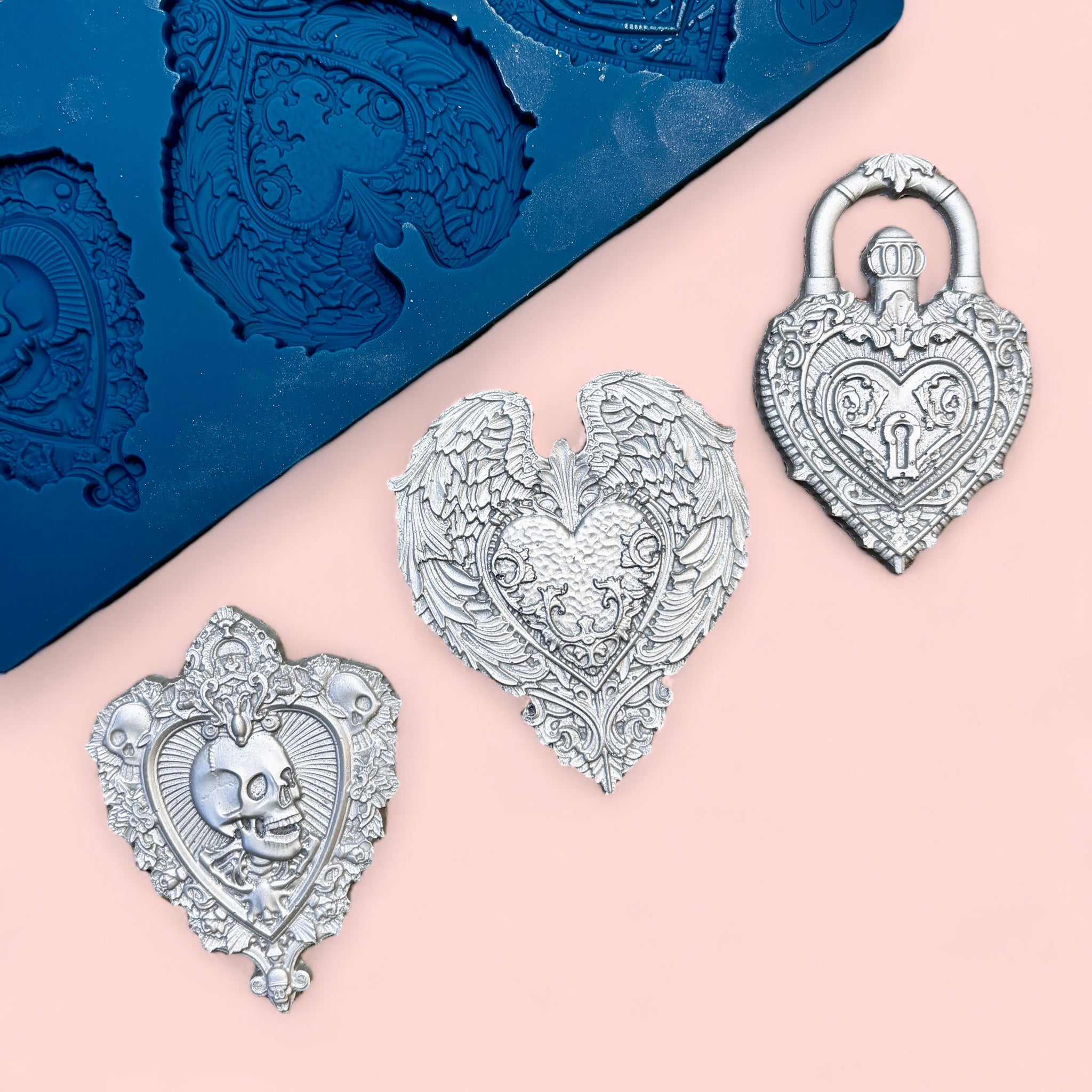 A blue silicone mold and silver colored castings of 3 hearts with ornate designs like skulls, wings, and a lock are against a light pink background.