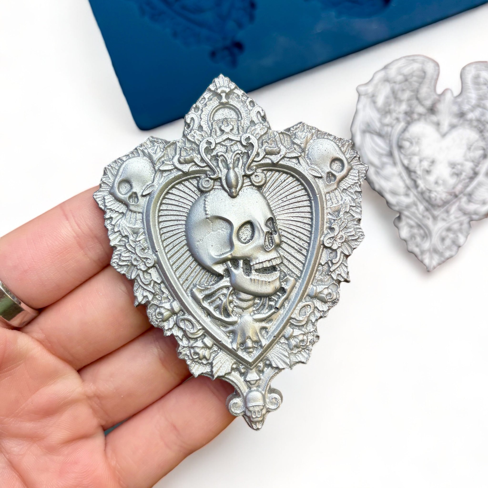 A blue silicone mold and silver colored castings of 3 hearts with ornate designs like skulls, wings, and a lock are against a white background. A hand is shown holding the heart with skulls.