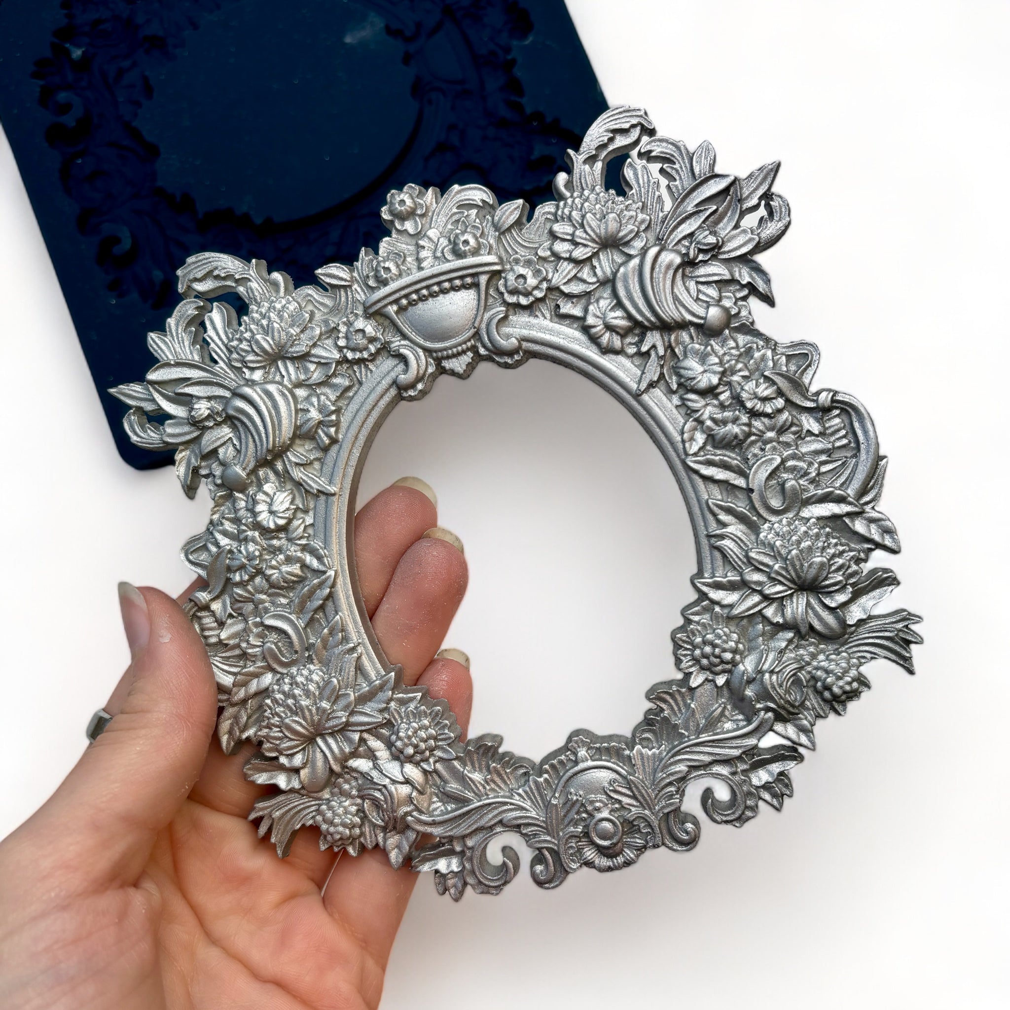 A blue silicone mold and siler colored castings of Zuri Design's Oval Floral Frame are against a white background. A hand is shown holding the frame casting.