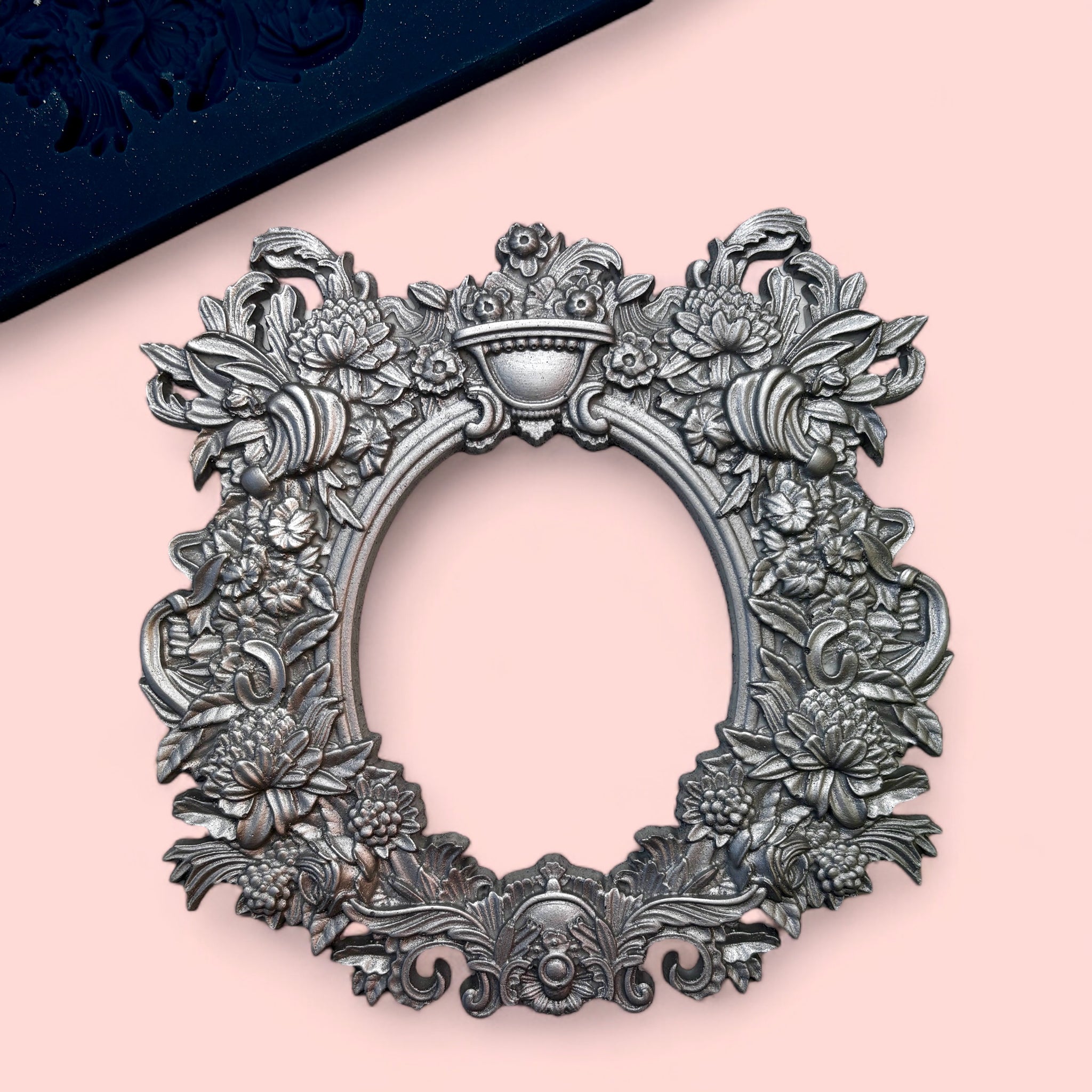 A blue silicone mold and silver colored casting of an oval ornately floral decorated frame is against a light pink background.