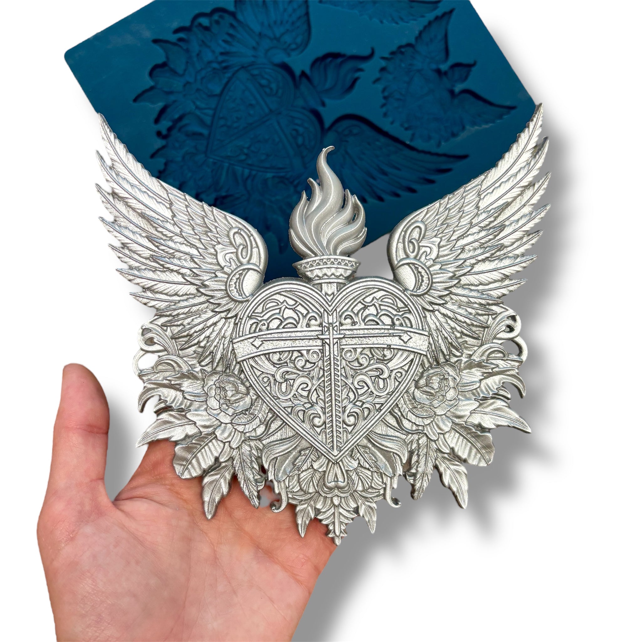 Zuri's Sacred Hearts silicone mold is against a white background. A hand is shown holding a silver colored casting of the large flame topped angel wing heart.