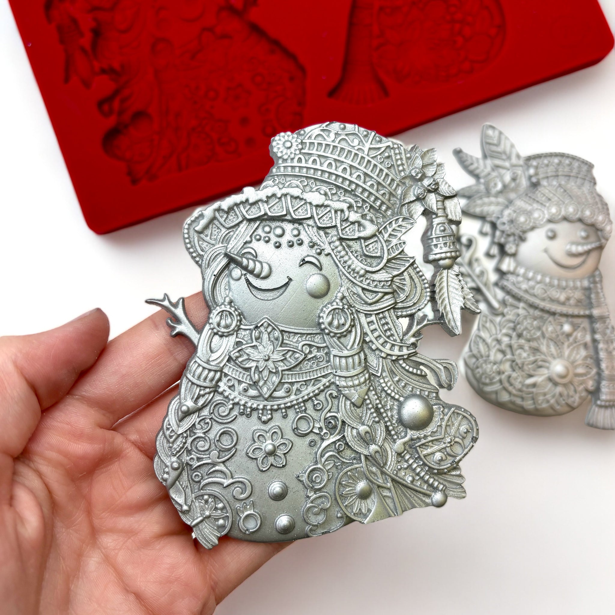 A red silicone mold and 2 silver colored silicone castings of 2 ornately decorated Bohemian style snowmen are against a white background. A hand is shown holding one of the castings.