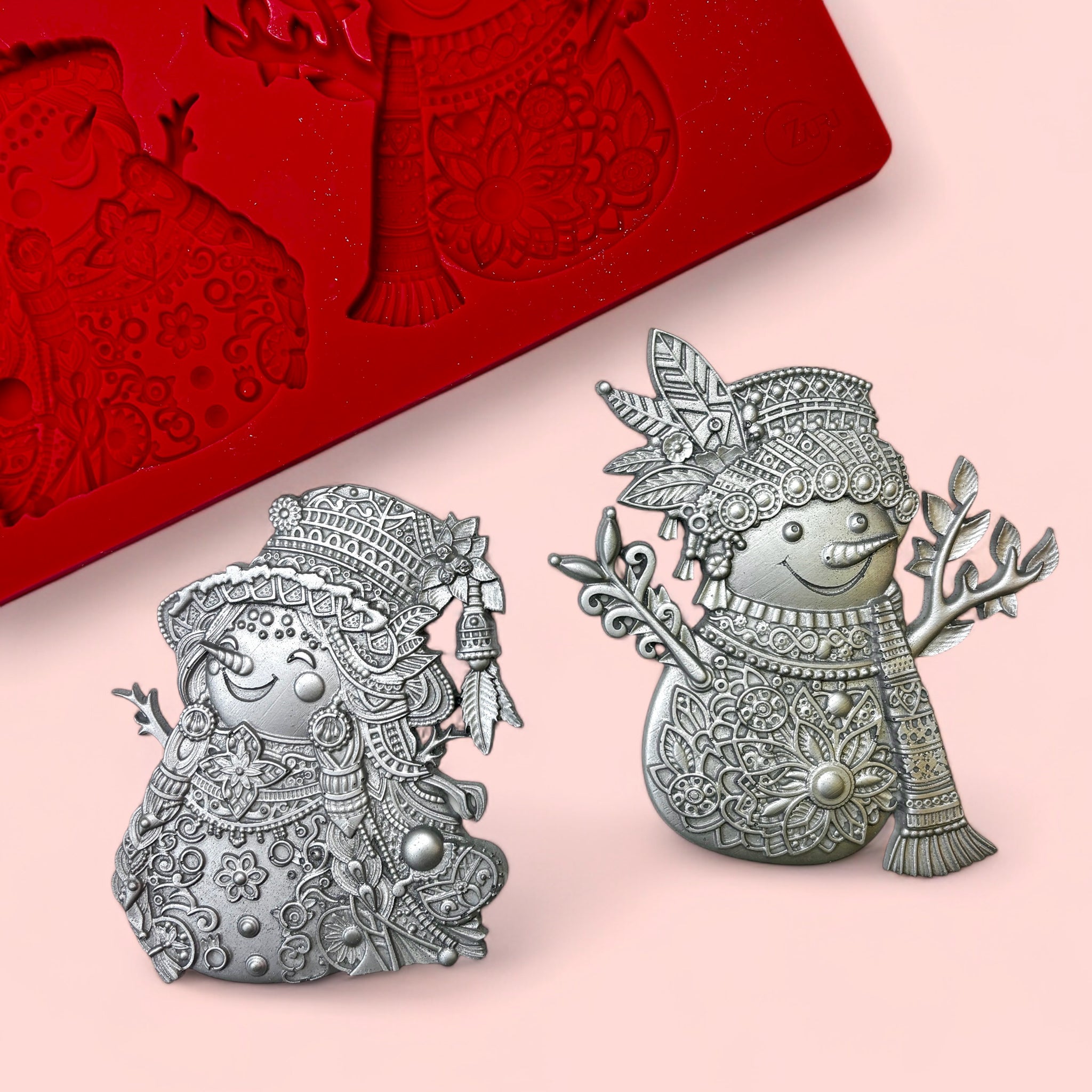 A red silicone mold and 2 silver colored silicone castings of 2 ornately decorated Bohemian style snowmen are against a light pink background.