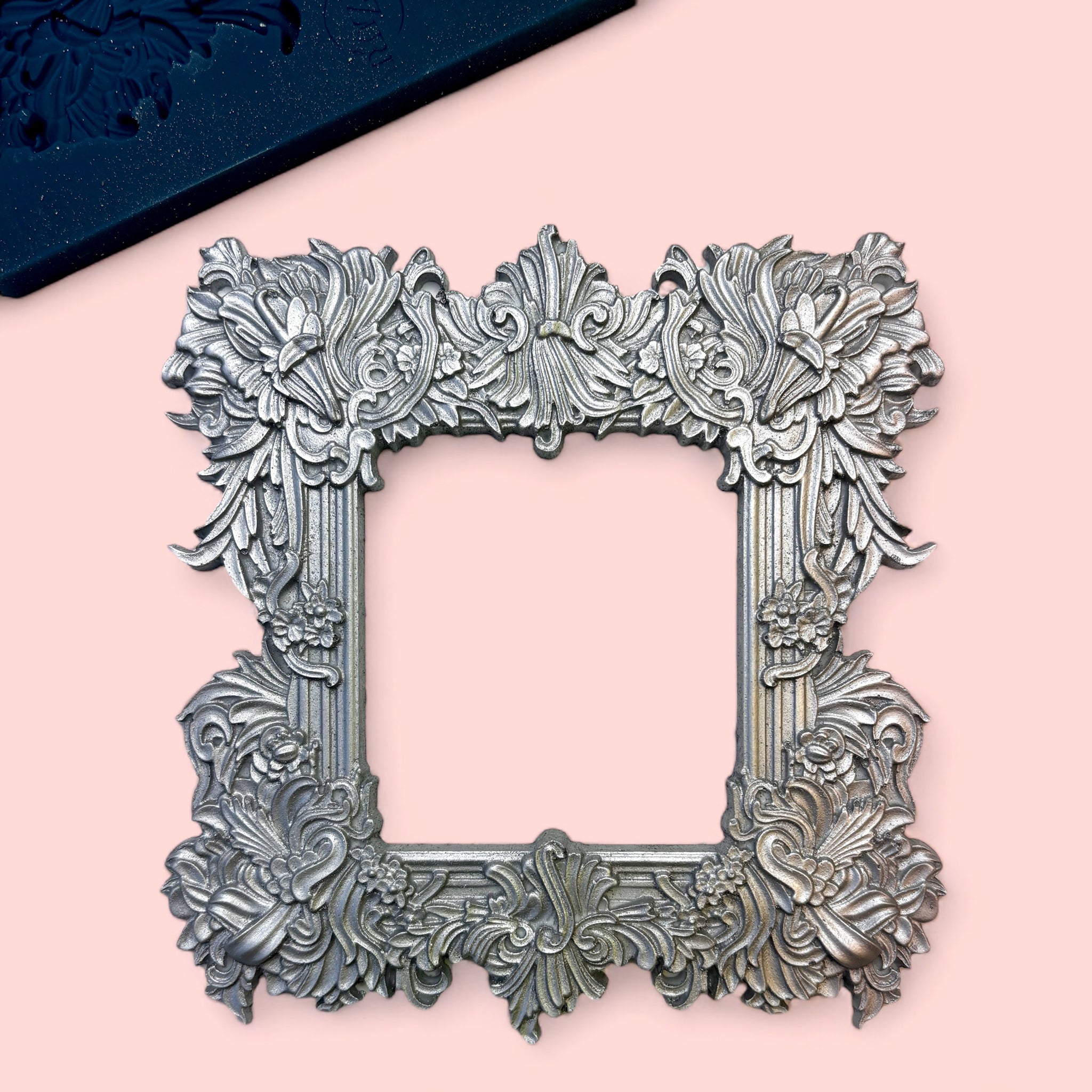 A blue silicone mold and silver colored casting of an ornate flourish square frame are against a light pink background.