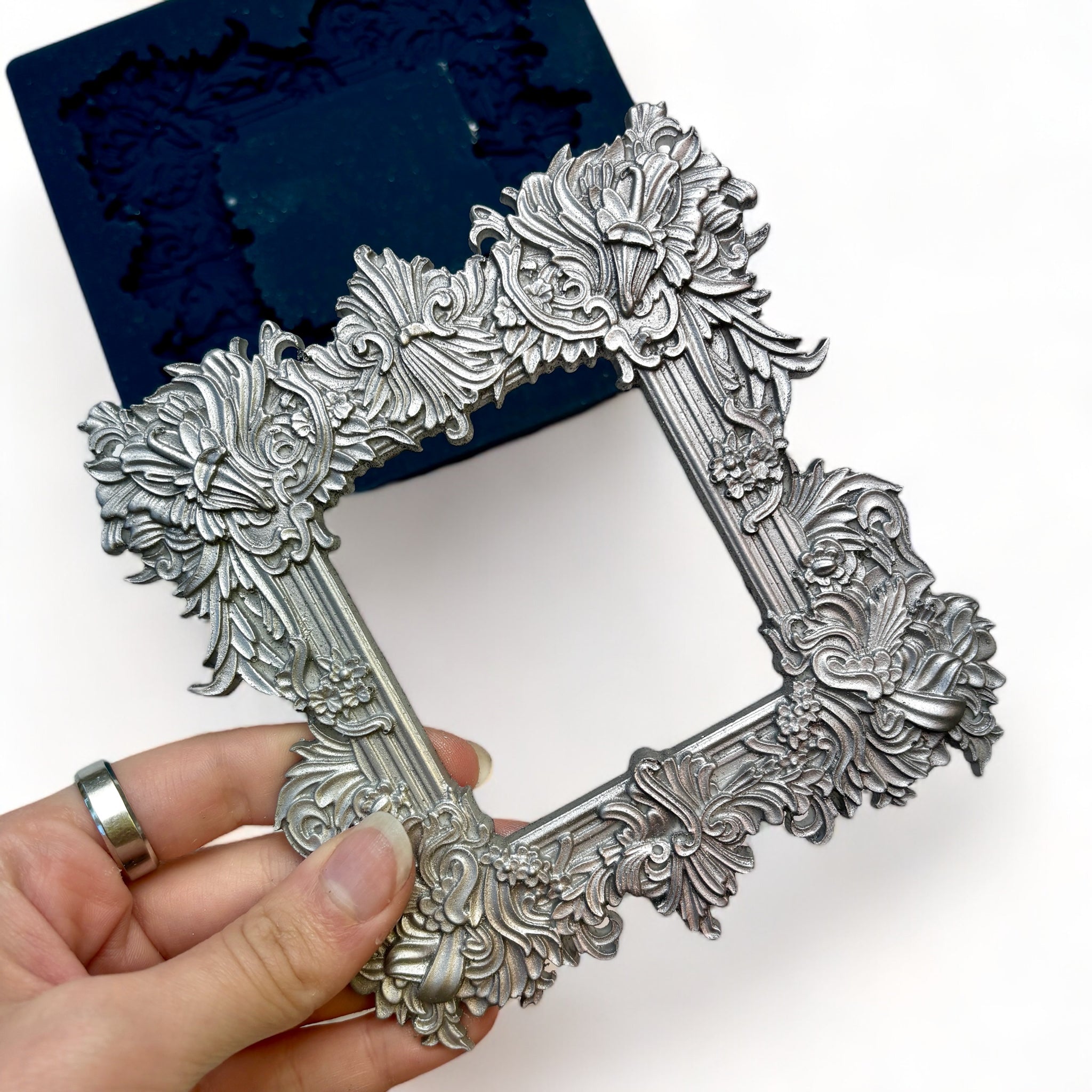 A blue silicone mold and silver colored casting of Zuri Design's Square Frame are against a white background. A hand is shown holding the frame casting.