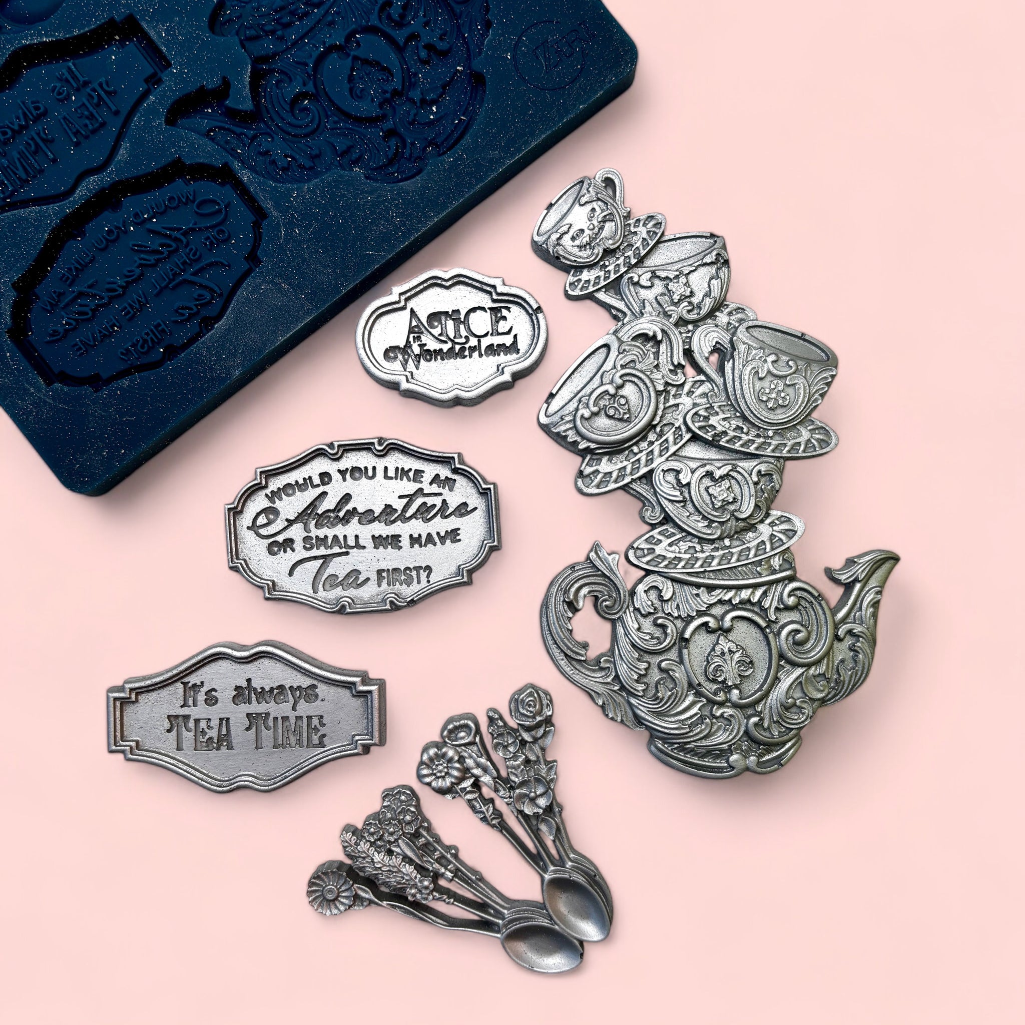 A blue silicone mold and silver colored castings of a stack of teacups on a tea pot, a cluster of floral handle spoons, and 3 small signs with Alice in Wonderland quotes are against a light pink background.