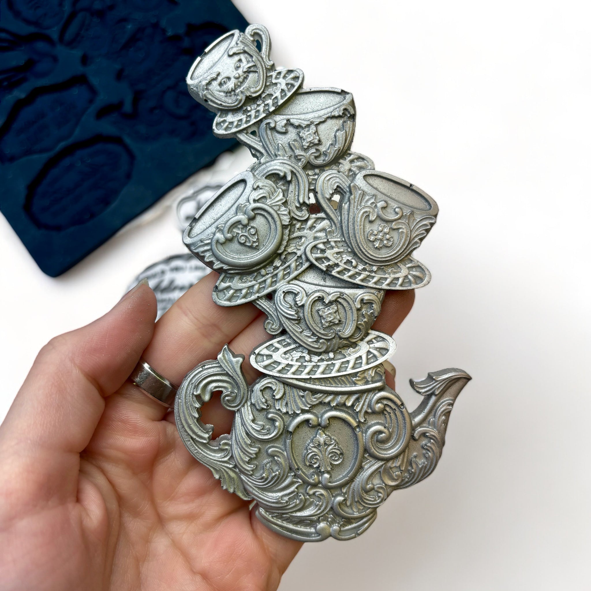 A blue silicone mold and silver colored castings of Zuri Design's Tea Time are against a white background. A hand is shown holding the stack of teacups on a teapot casting.