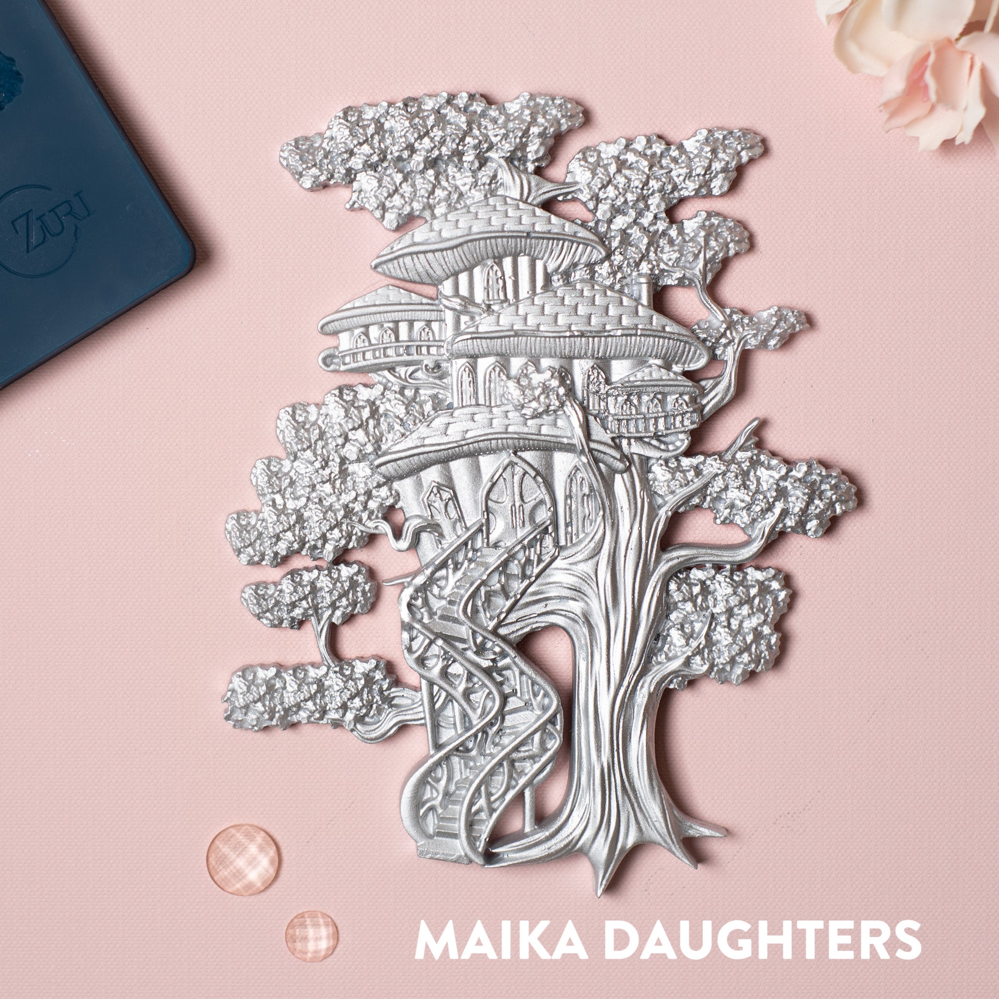 A blue silicone mold and silver colored casting of a tree house  made of mushrooms are against a light pink background.