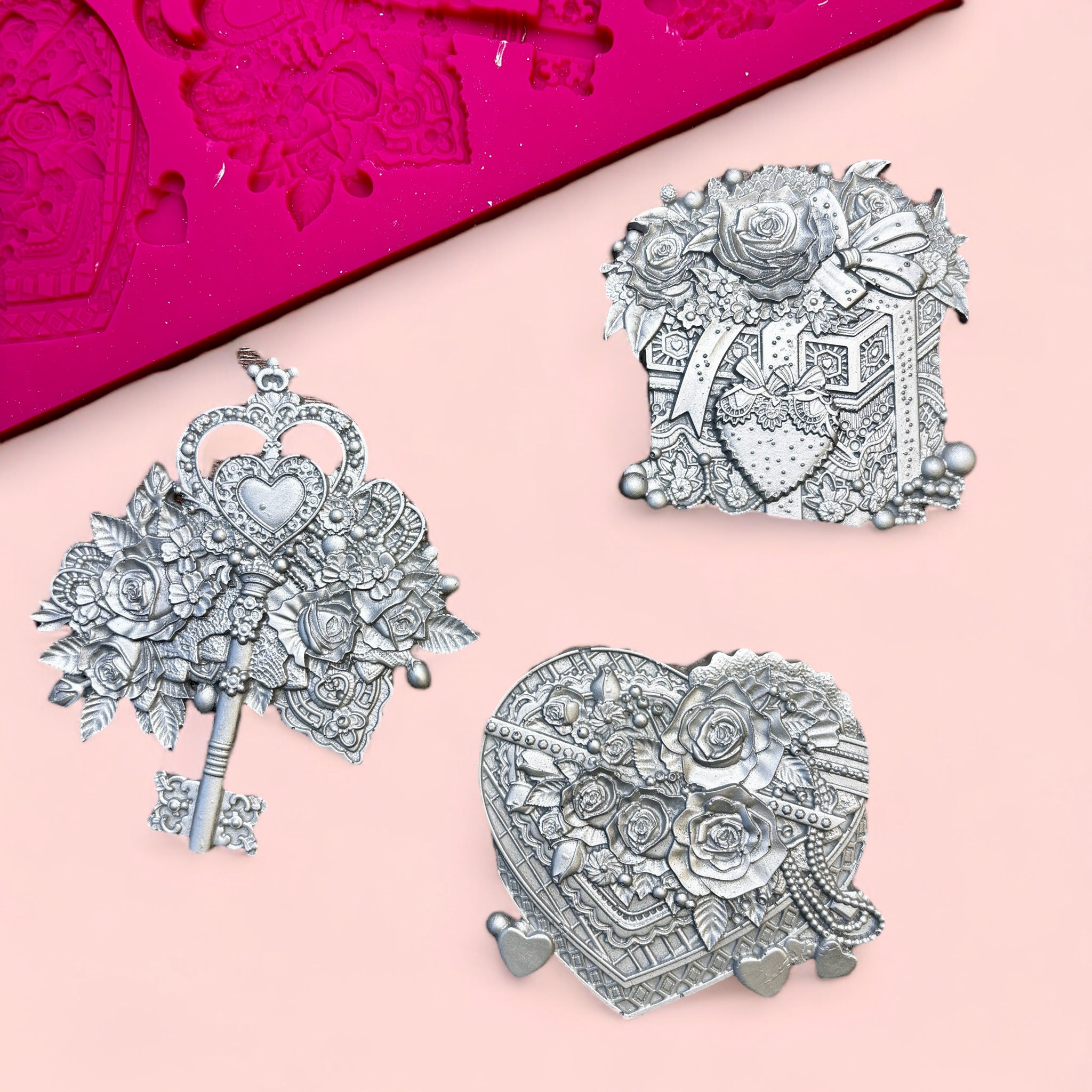 A pink silicone mold and silver colored castings of a Valentine's Day gift box, box of chocolates, and a key surrounded by flowers are against a light pink background.