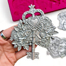 A pink silicone mold and silver colored castings of a Valentine's Day gift box, box of chocolates, and a key surrounded by flowers are against a white background. A hand is shown holding the key casting.