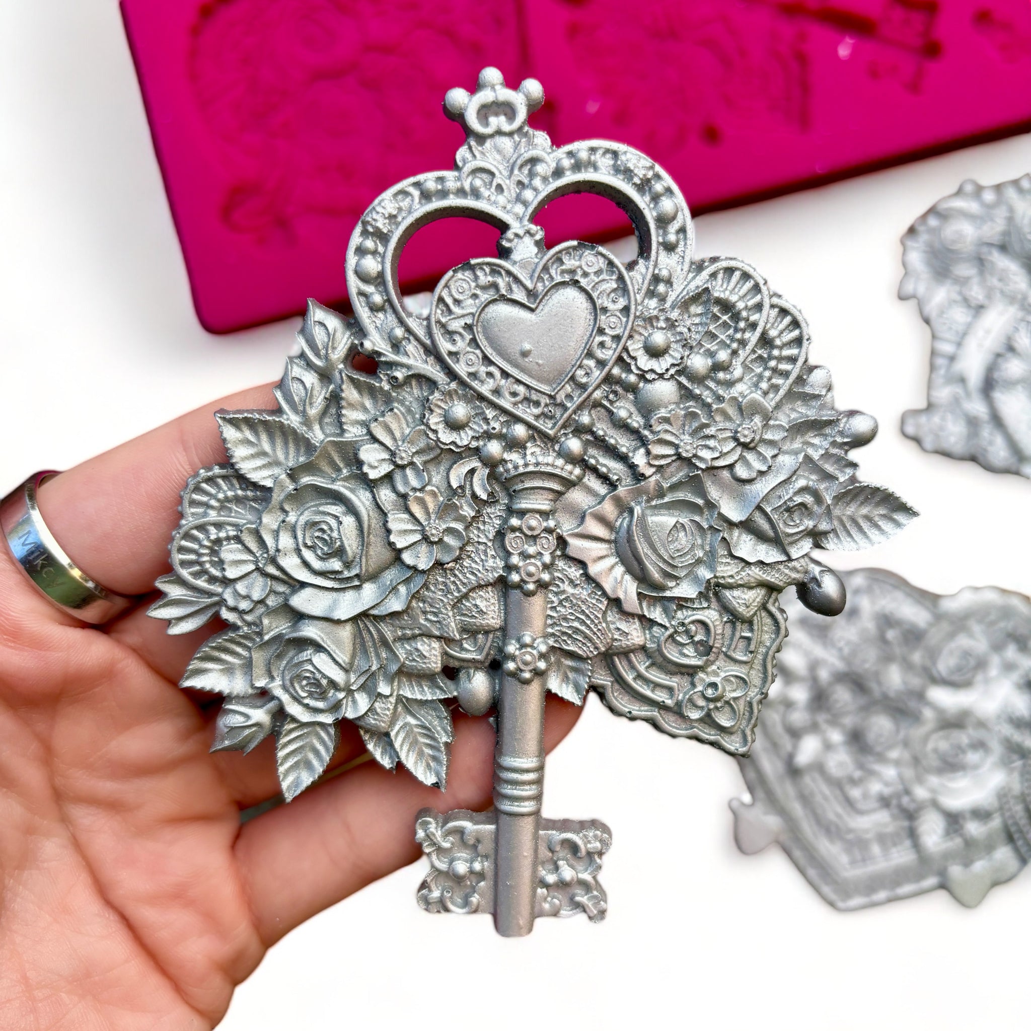 A pink silicone mold and silver colored castings of a Valentine's Day gift box, box of chocolates, and a key surrounded by flowers are against a white background. A hand is shown holding the key casting.