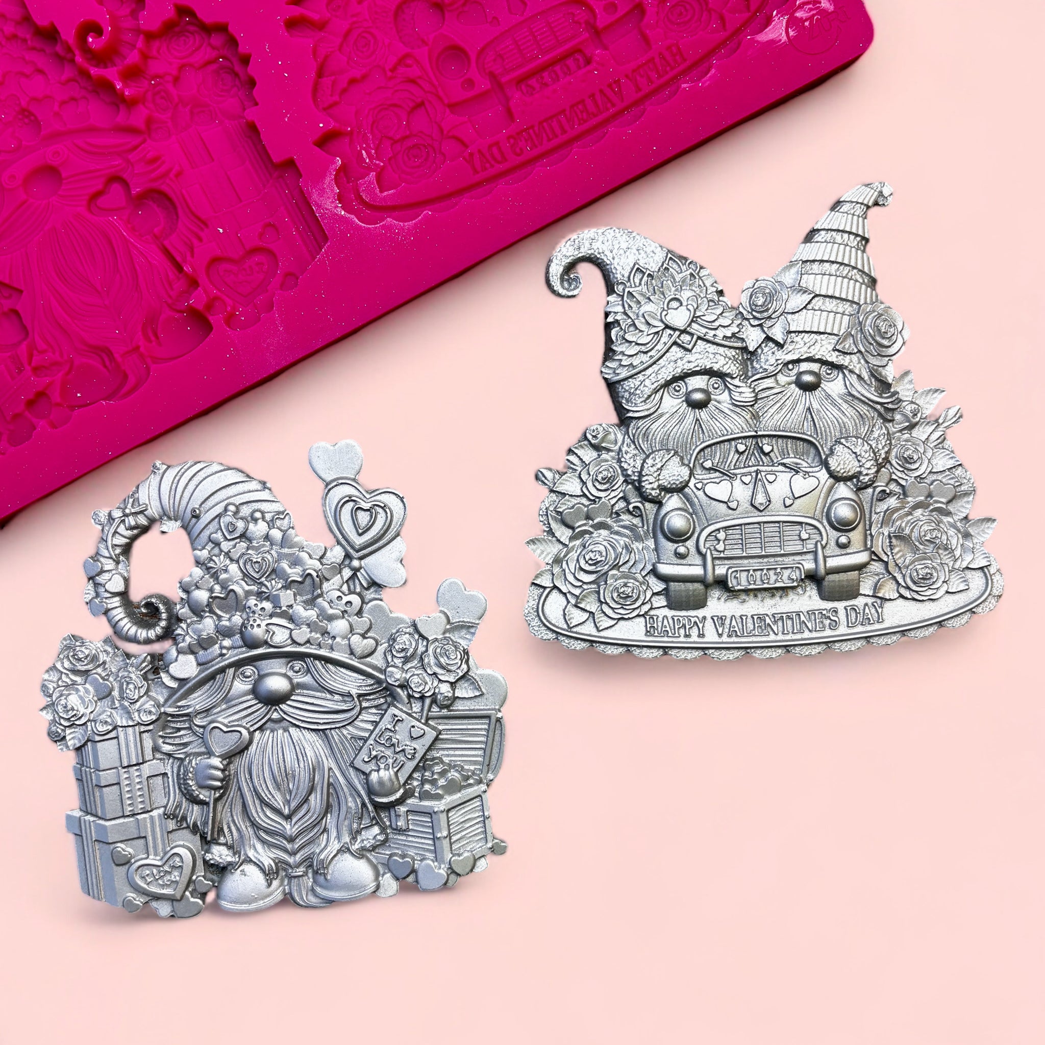 A pink silicone mold and 2 silver colored castings of Valentine's Day themed gnomes are against a light pink background. 