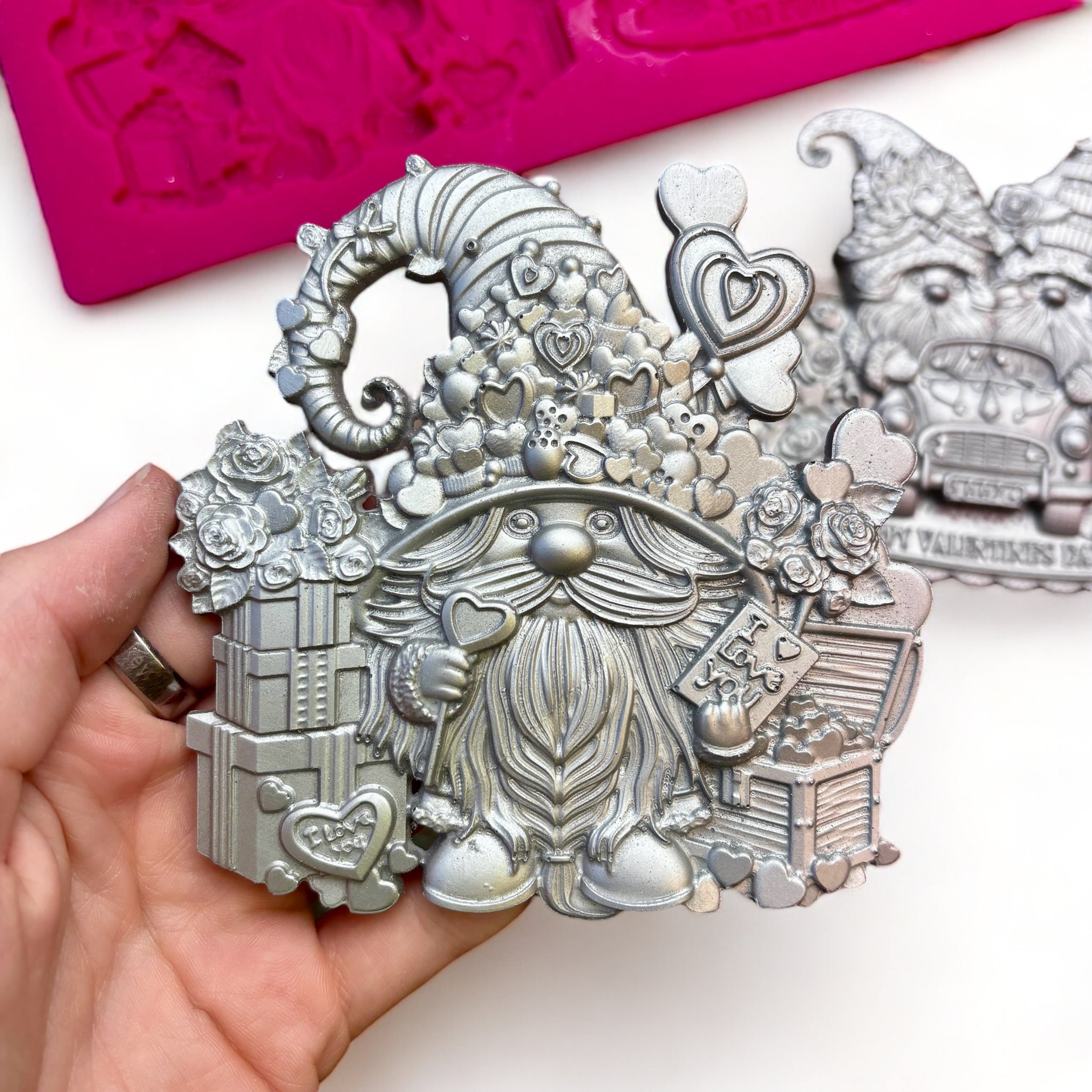 A pink silicone mold and 2 silver colored castings of Valentine's Day themed gnomes are against a white background. A hand is shown holding one of the castings.