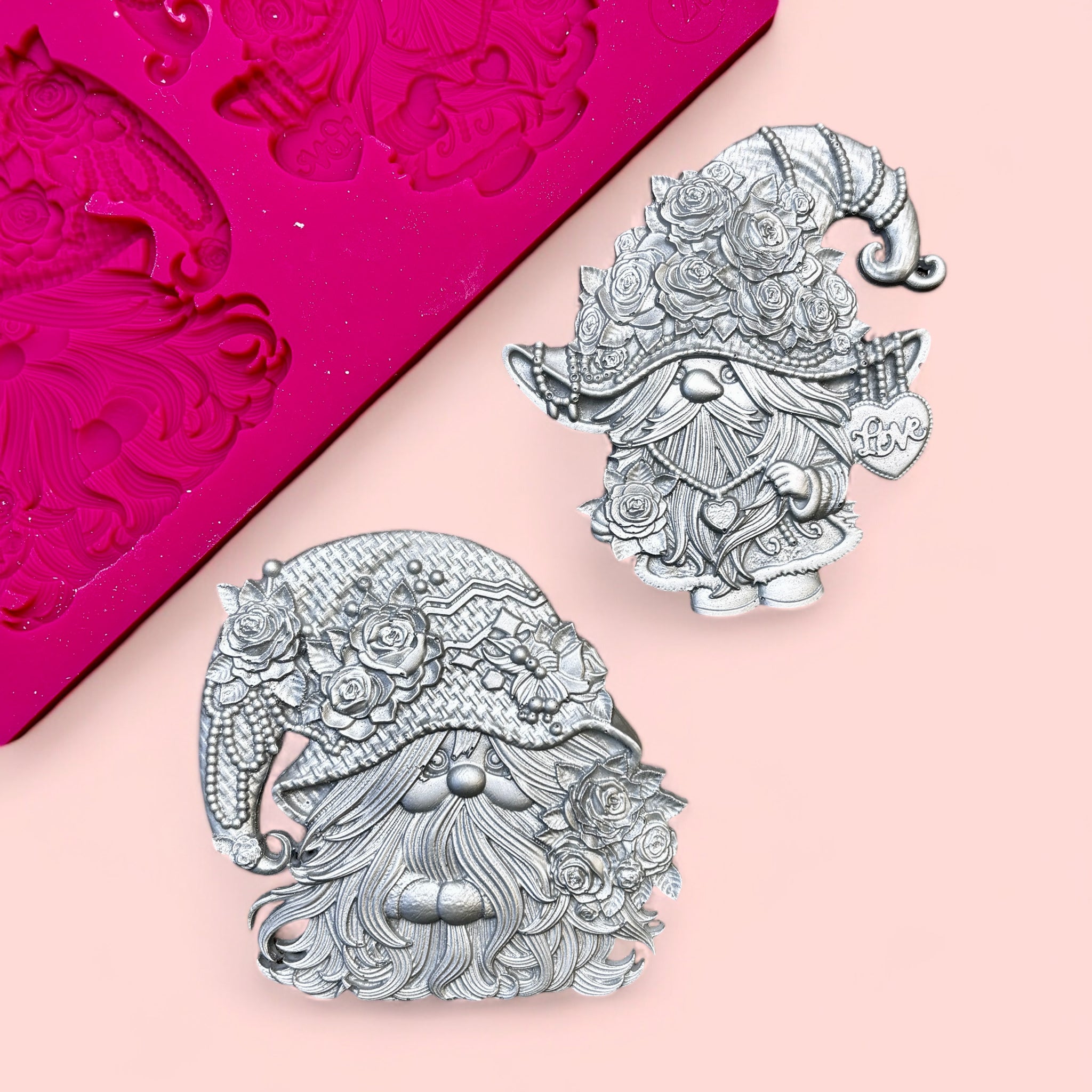 A pink silicone mold and 2 silver colored castings of Valentine's Day themed gnomes are against a light pink background.