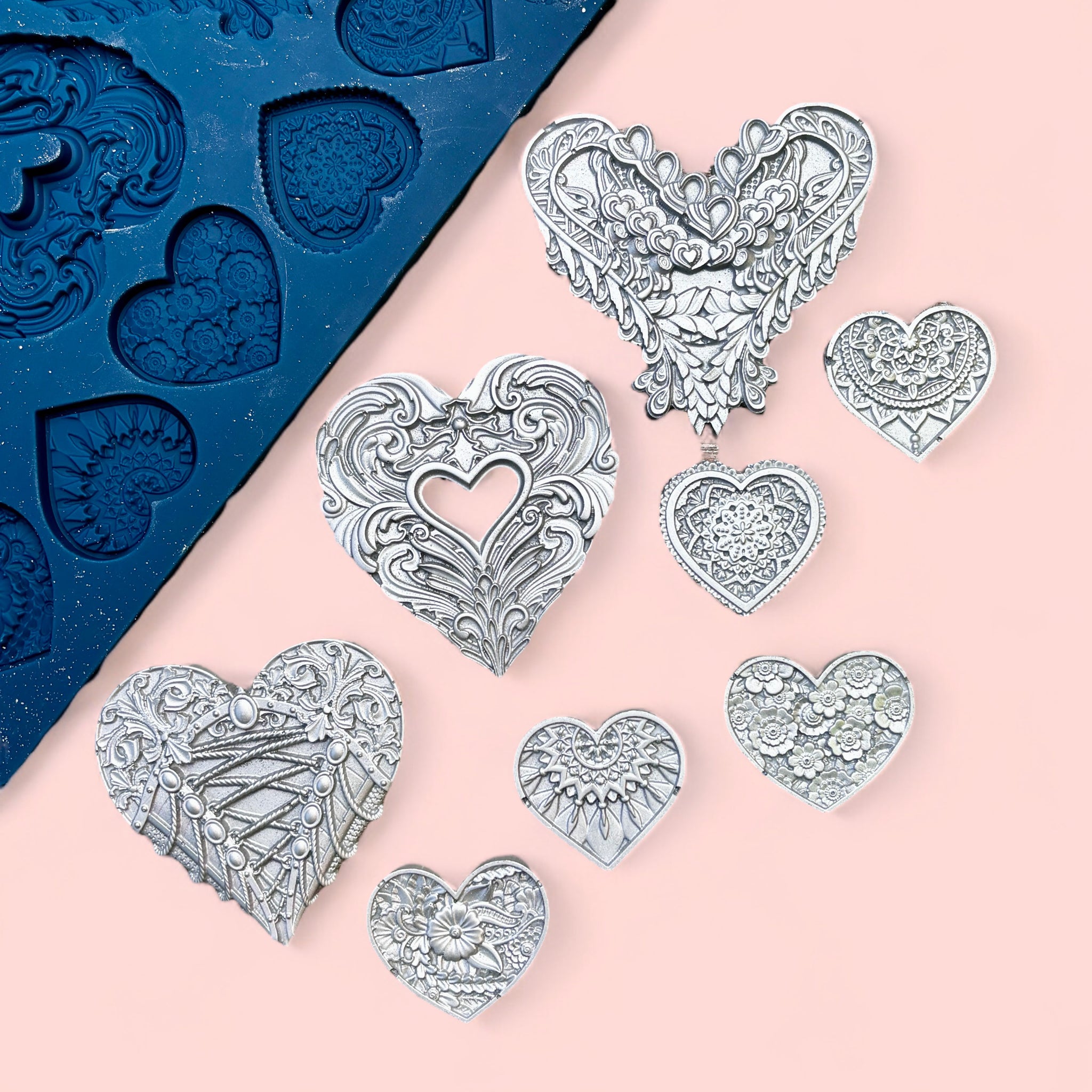 A blue silicone mold and silver colored castings of 3 large and 5 small ornately decorated hearts are against a light pink background.