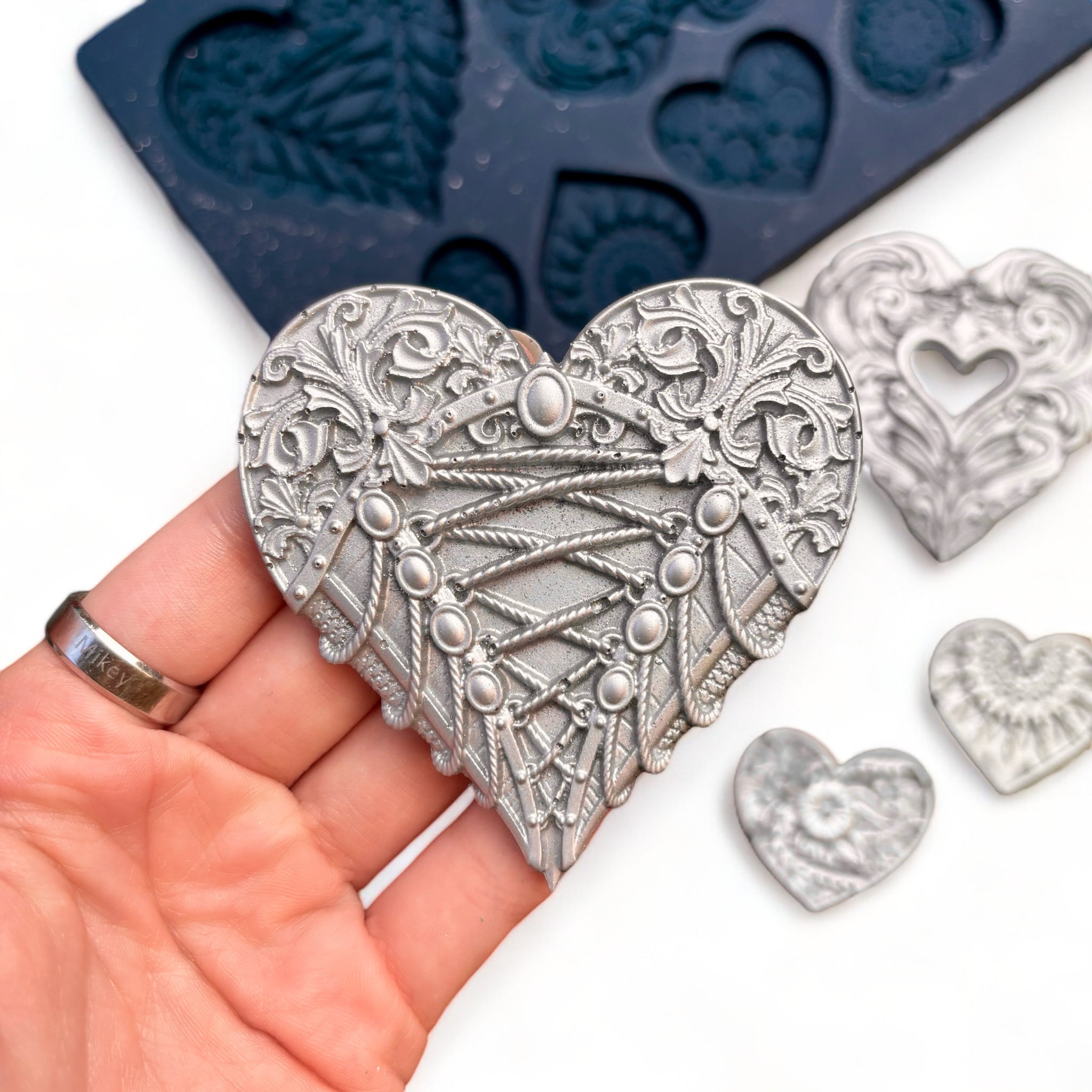 A blue silicone mold and silver colored castings of 3 large and 5 small ornately decorated hearts are against a white background. A hand is shown holding one of the large heart castings.