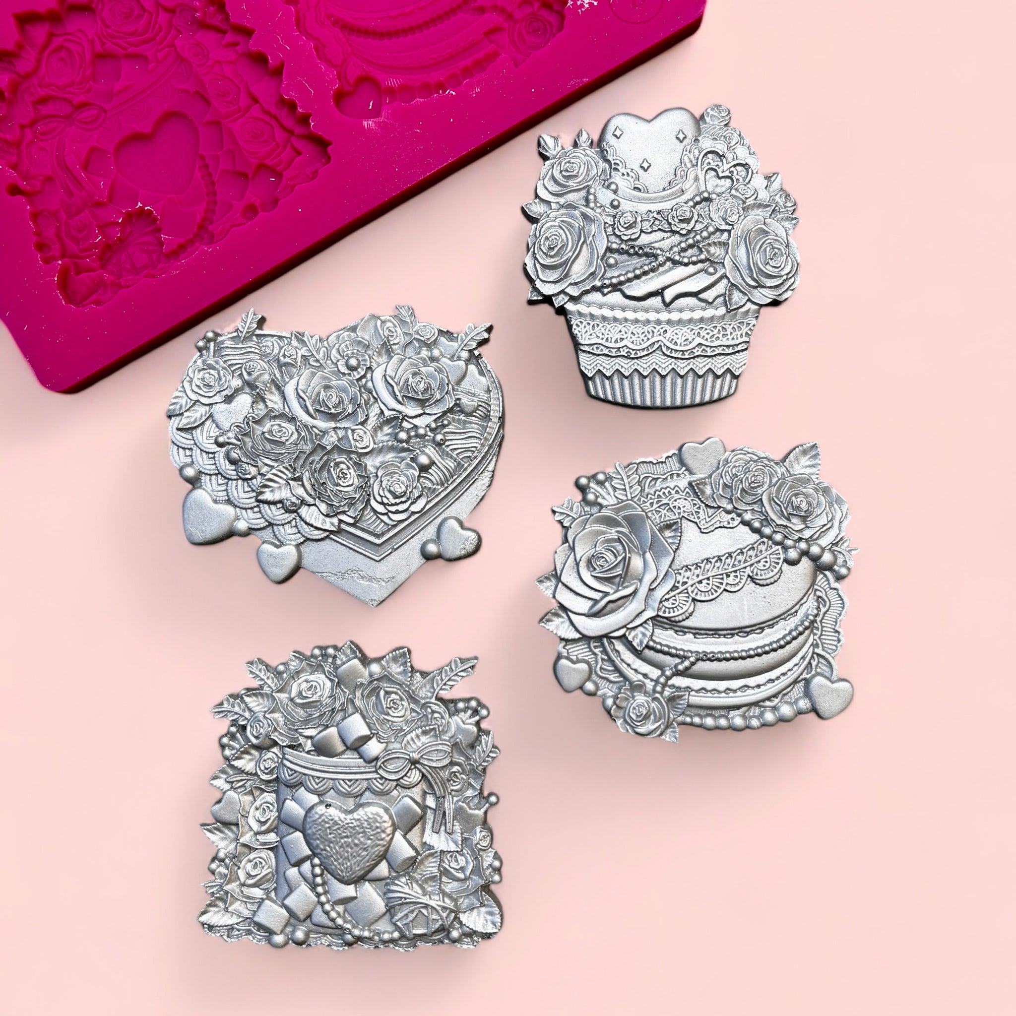 A pink silicone mold and silver colored castings of 4 Valentine's Day candy boxes surrounded with flowers are against a light pink background.